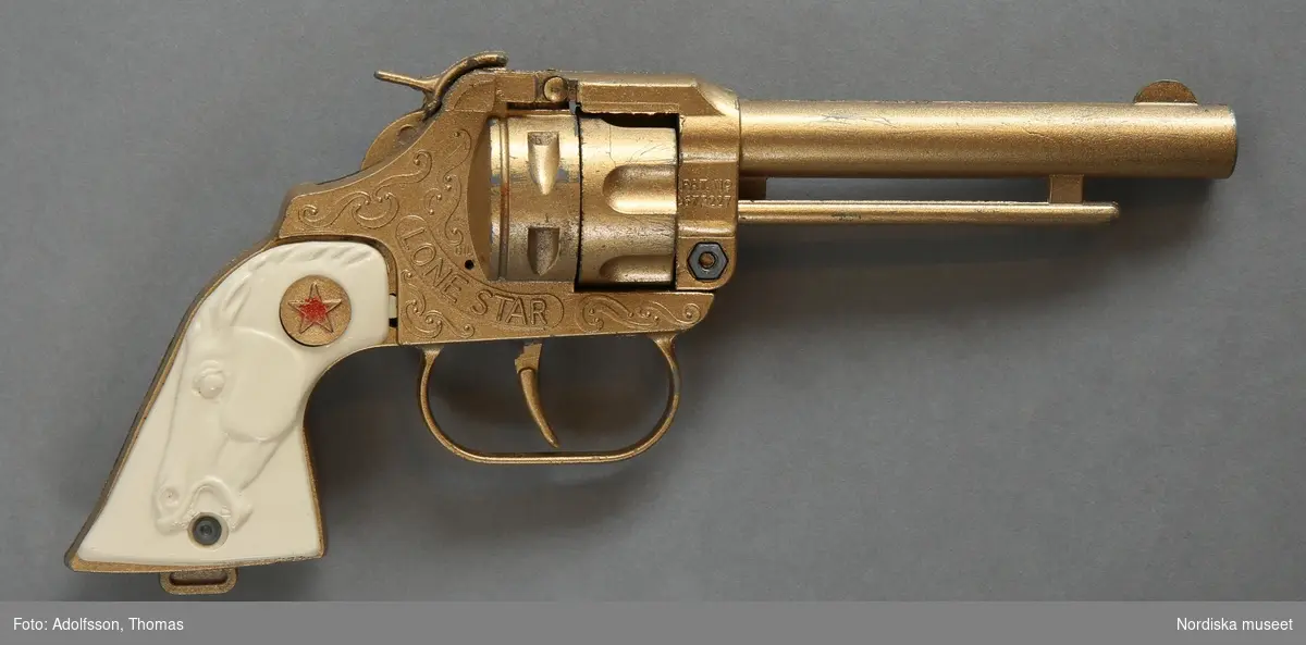 Inventering Sesam 1996-1999:
Revolver L 21 cm, B 2 cm, H 7,5 cm
Bälte L 81cm, B 4 cm Hölster L 24 cm.
Patroner 3,5 cm, Diam 1 cm
Revolver med bälte, hölster och 3 patroner
Revolver: gjuten i metall i två delar. Gulbronserad. Slät pipa, stock med dekorslingor, varbygel vid avtryckaren, kolven täckt på sidorna av vit plast med hästhuvud. Vid laddning spänner man ut hanen och tar bort spärren på höger sida, då öppnas pistolen och man kan ladda med en rulle knallpulver som fästs på den lilla piggen bakom cylindern och läggs i banan bakom piggen, så att när man trycker på avtryckaren slår hanen på knallpulverremsan. När man skjuter skall knallpulverremsan matas upp automatiskt vid hanen. 
Revolvern märkt: på sidorna av stocken "LONE STAR". Mellan pipa och stock "PAT. NO 677227". Vid cylindern (syns när den är öppen) "DCMT / LTD / SLIKKA TOY / MADE IN / ENGLAND" DCMT står för Diecast Casting Machine Tools Ltd. Se vidare "Millers Toys and Games Aniques Checklist" av Hugo Marsh, London 1995, sid 124.
Bälte och Hölster: av läder, grön och vit. Spänne av metall. Hölster fäst vid bältet med nitar, hölstret dekorerat med nitar i rött, blått och silver. Ovanför hölstret tre hållare i läder för patroner.
Patroner: tre stycken av trä, gulmålade, spetsen i silver.
Leif Wallin 1996