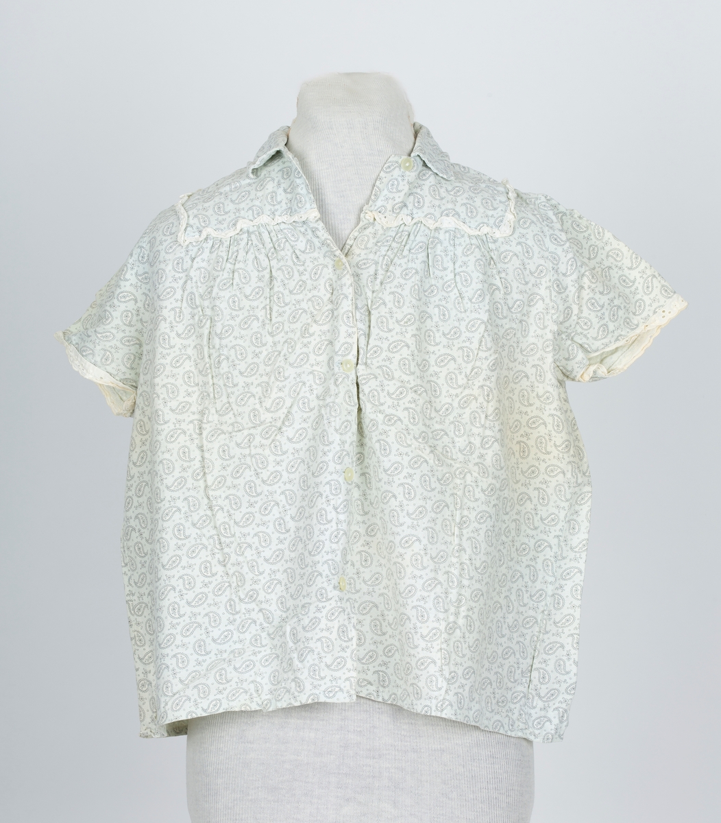 Pyjamas, fllanellignende stogg påtrykt dråpemønster med blomst inni. 

A: Overdel 
Forknappet, 5 knapper og knapphull. Krage. Firkantet bærestykke kantet med maskinlaget engelsk blonde. Kort erme kantet med engelsk blonde.
B: Bukse
Knebukse. Løpegang for strikk i livet. Engelsk blondekant på bukseben.