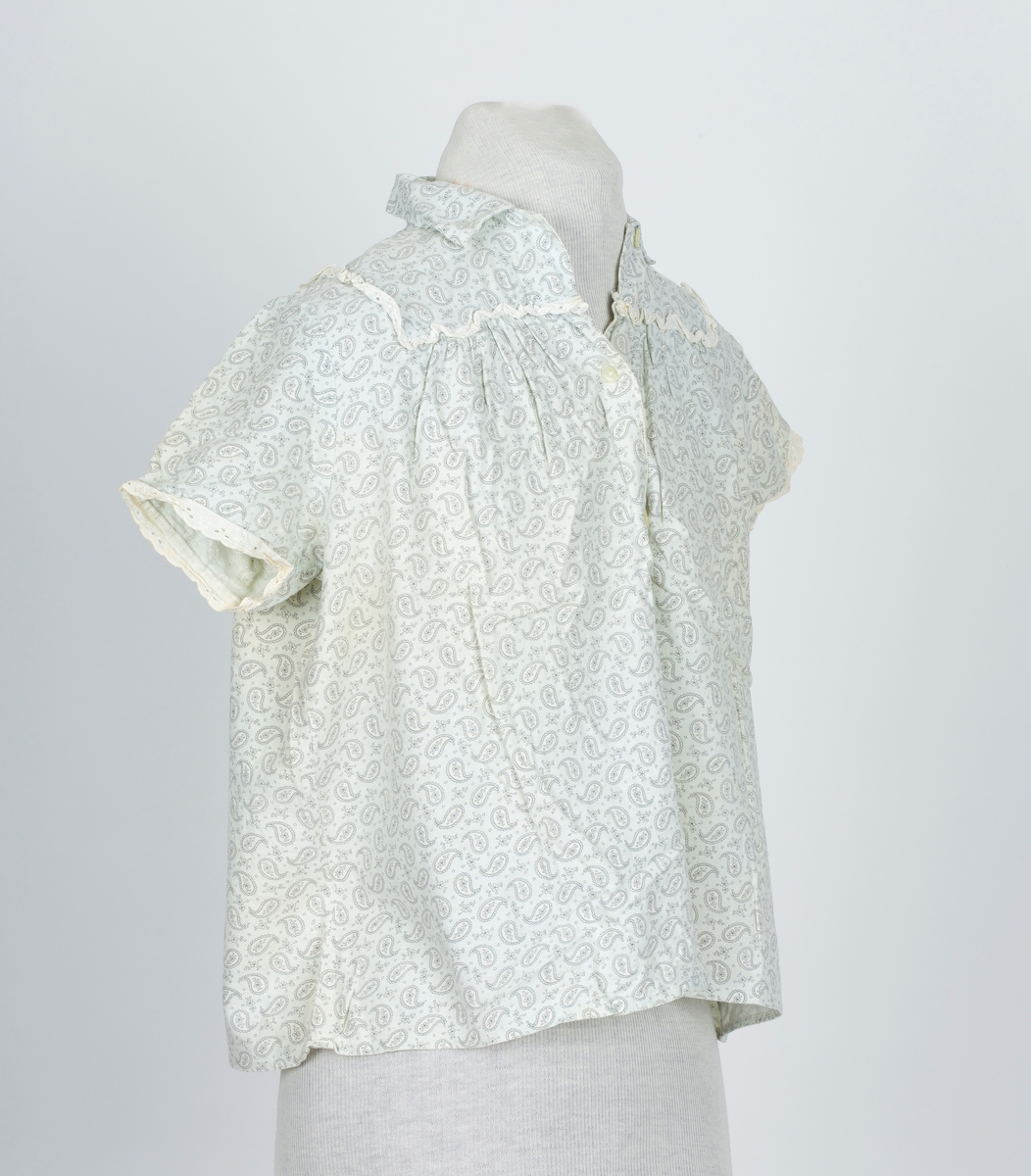 Pyjamas, fllanellignende stogg påtrykt dråpemønster med blomst inni. 

A: Overdel 
Forknappet, 5 knapper og knapphull. Krage. Firkantet bærestykke kantet med maskinlaget engelsk blonde. Kort erme kantet med engelsk blonde.
B: Bukse
Knebukse. Løpegang for strikk i livet. Engelsk blondekant på bukseben.