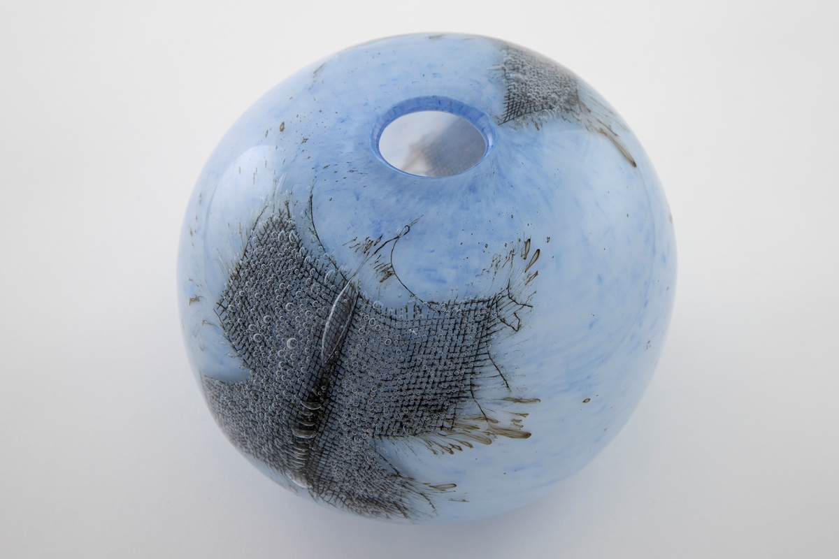 Kuleformet vase med liten rund munning i overfangsglass. Halvgjennomskinnelig melkehvitt glass med lyseblå fargepigmenter samt to innlagte metallnett. Sistnevnte danner et kryss, og ut ifra disse stråler lange, spisse luftbobler.