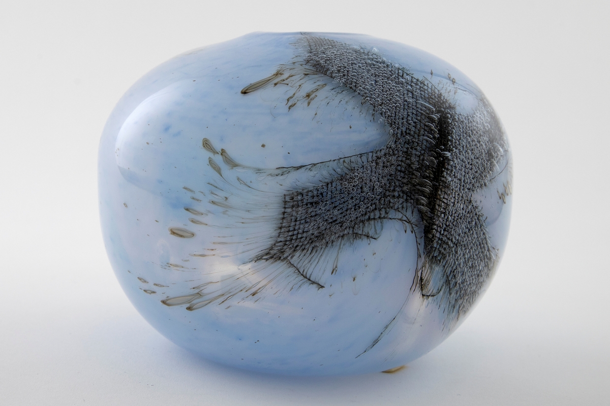 Kuleformet vase med liten rund munning i overfangsglass. Halvgjennomskinnelig melkehvitt glass med lyseblå fargepigmenter samt to innlagte metallnett. Sistnevnte danner et kryss, og ut ifra disse stråler lange, spisse luftbobler.