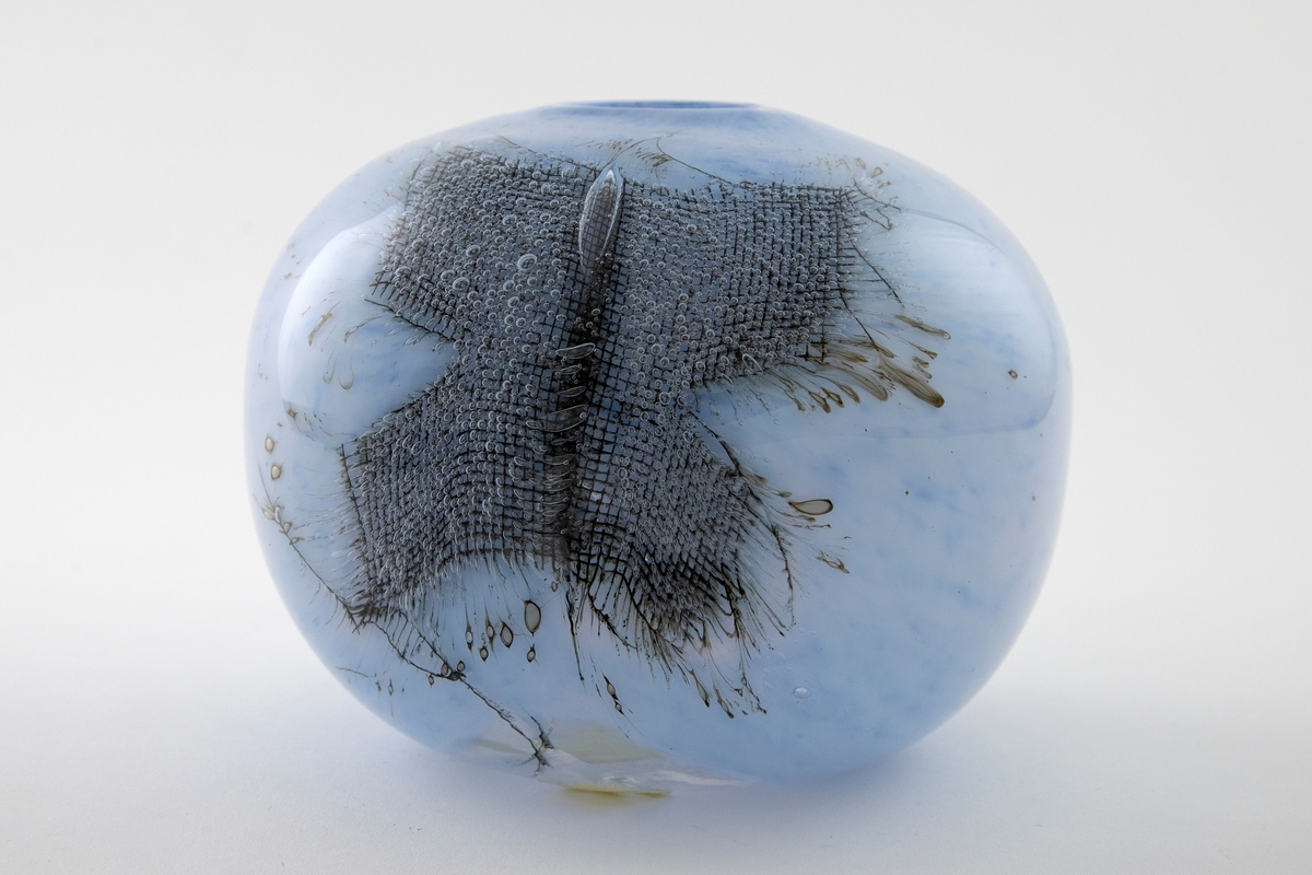 Kuleformet vase med liten rund munning i overfangsglass. Halvgjennomskinnelig melkehvitt glass med lyseblå fargepigmenter samt to innlagte metallnett. Sistnevnte danner et kryss, og ut ifra disse stråler lange, spisse luftbobler.