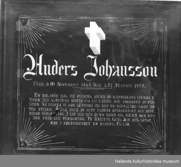 Begravningstavla målad på glas. a) Svart bakgrund. Den dödes namn samt ett kors ovanför i stanniolpapper, (fästat på baksidan). I varje hörn en sol som strålar mot centrum. b) Ram, bredd 3,5 cm. Ram av trä, profilerad. Svartmålad.Text: "Anders Johansson Född d. 10 November 1862"