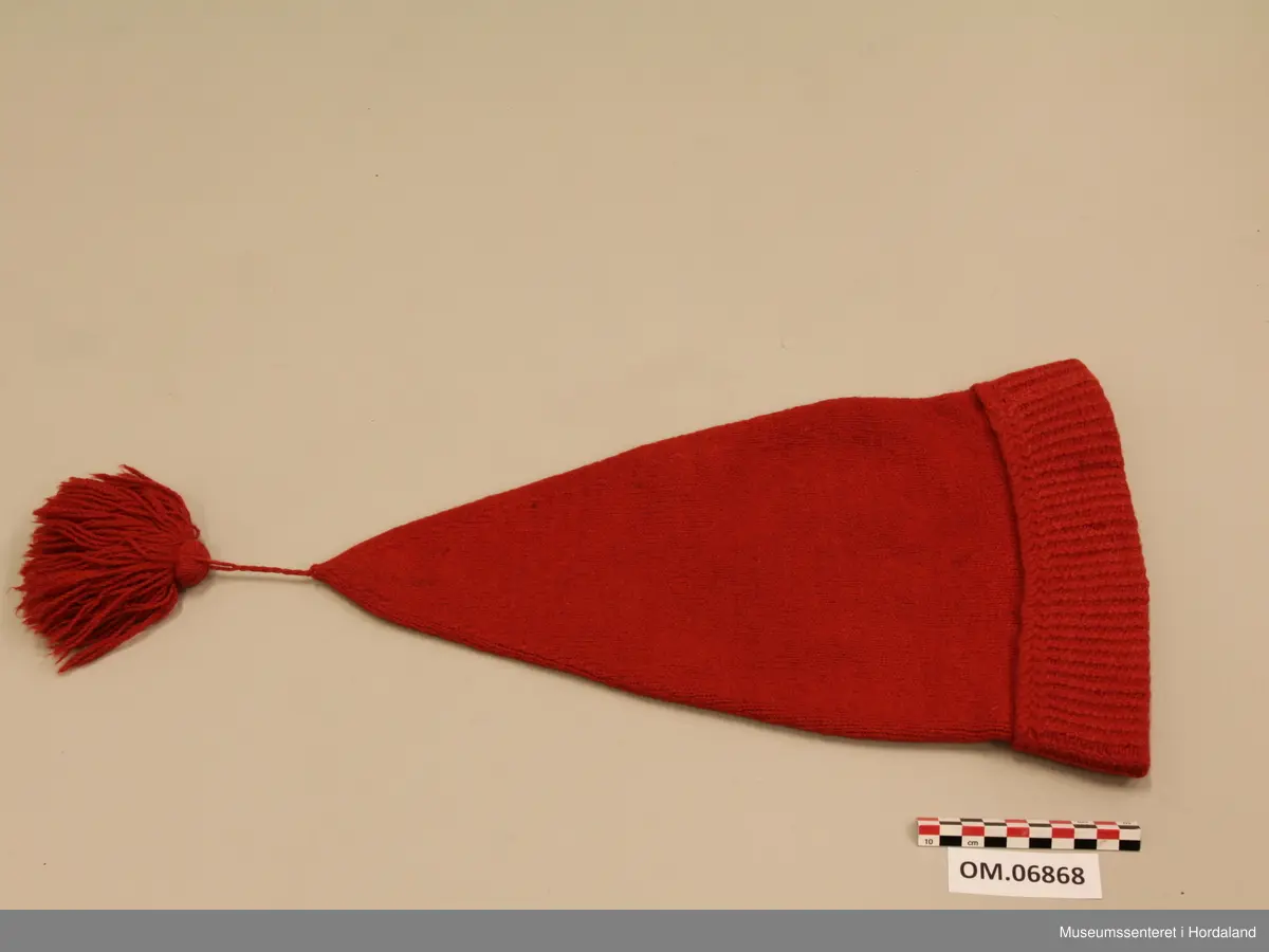 Raud ullhue med dusk. Lang modell som spissar jamt mot toppen. Oppbretta kant med mønsterstrikk. Tvinna snor i toppen, og fyldig frynsedusk.