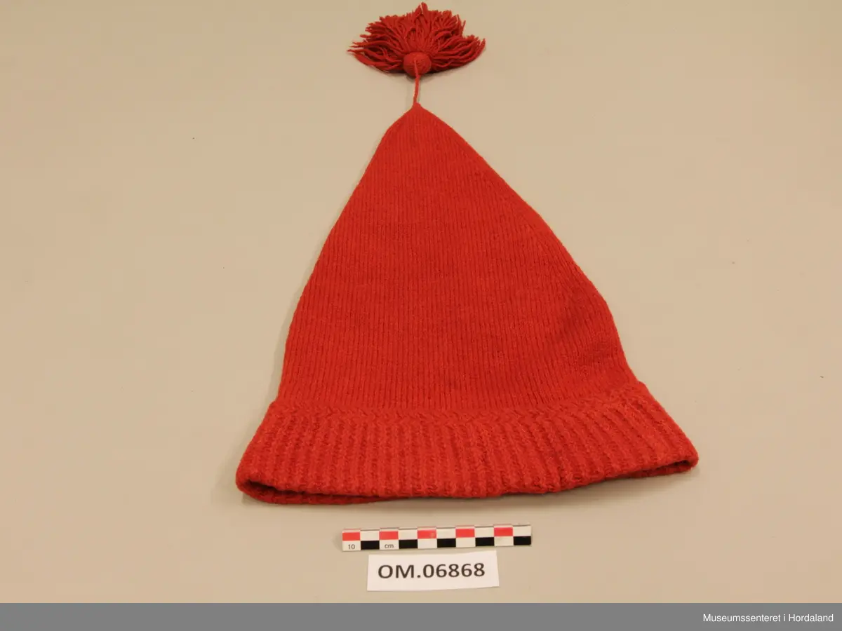 Raud ullhue med dusk. Lang modell som spissar jamt mot toppen. Oppbretta kant med mønsterstrikk. Tvinna snor i toppen, og fyldig frynsedusk.