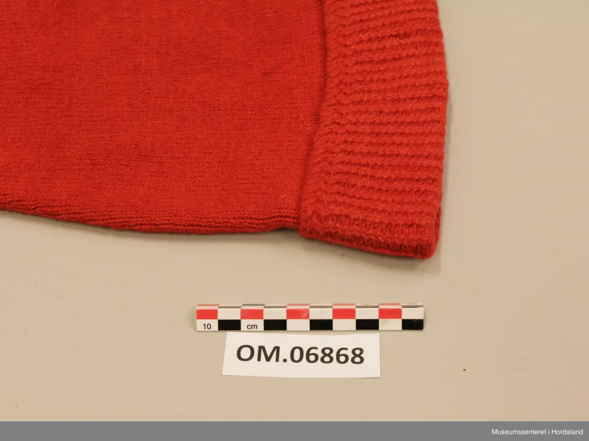 Raud ullhue med dusk. Lang modell som spissar jamt mot toppen. Oppbretta kant med mønsterstrikk. Tvinna snor i toppen, og fyldig frynsedusk.