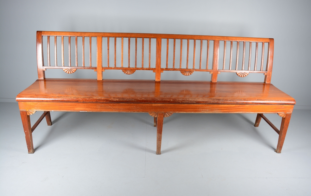 Sittebenk som har stått i Svaneapotekets kundeareal. Plass til 4 personer. Svakt buet ryggdel som danner ryggdel og ben på benken i samme stykke. Ryggen er delt i fire felt med lik dekor i hvert felt, 7 loddrette staver inni et rektangulært felt. En halvmåneformet blomst på midten av hvert felt på undersiden av det rektangulære feltet. Setet er laget i ett stykke tre og har en svak krumming oppover frem mot kanten på benken. Benken har tre sett med ben, to og to, på endene og på midten. Bena er parvis festet sammen med en sidesprosse. Rette framben. Under sargen på sidene og fremme er benken dekorert med samme type blomst som på ryggdelen, men her er de en halv halvmåne.