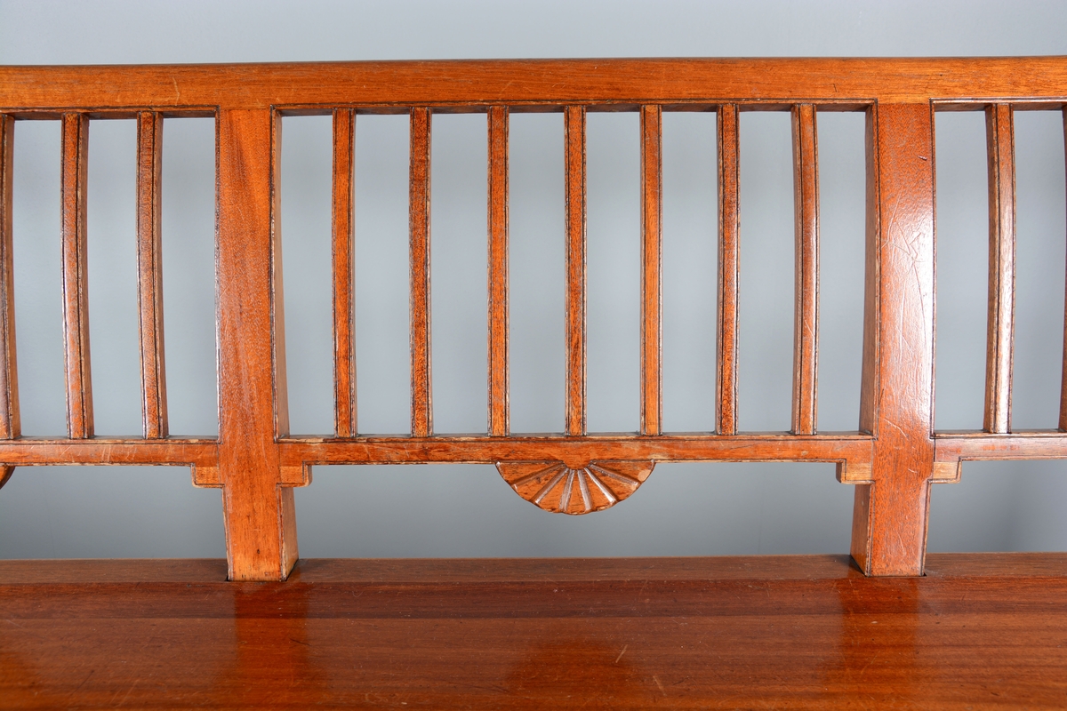 Sittebenk som har stått i Svaneapotekets kundeareal. Plass til 4 personer. Svakt buet ryggdel som danner ryggdel og ben på benken i samme stykke. Ryggen er delt i fire felt med lik dekor i hvert felt, 7 loddrette staver inni et rektangulært felt. En halvmåneformet blomst på midten av hvert felt på undersiden av det rektangulære feltet. Setet er laget i ett stykke tre og har en svak krumming oppover frem mot kanten på benken. Benken har tre sett med ben, to og to, på endene og på midten. Bena er parvis festet sammen med en sidesprosse. Rette framben. Under sargen på sidene og fremme er benken dekorert med samme type blomst som på ryggdelen, men her er de en halv halvmåne.