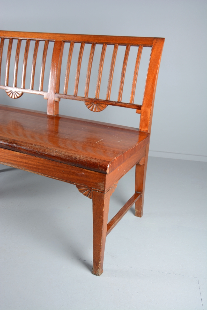 Sittebenk som har stått i Svaneapotekets kundeareal. Plass til 4 personer. Svakt buet ryggdel som danner ryggdel og ben på benken i samme stykke. Ryggen er delt i fire felt med lik dekor i hvert felt, 7 loddrette staver inni et rektangulært felt. En halvmåneformet blomst på midten av hvert felt på undersiden av det rektangulære feltet. Setet er laget i ett stykke tre og har en svak krumming oppover frem mot kanten på benken. Benken har tre sett med ben, to og to, på endene og på midten. Bena er parvis festet sammen med en sidesprosse. Rette framben. Under sargen på sidene og fremme er benken dekorert med samme type blomst som på ryggdelen, men her er de en halv halvmåne.