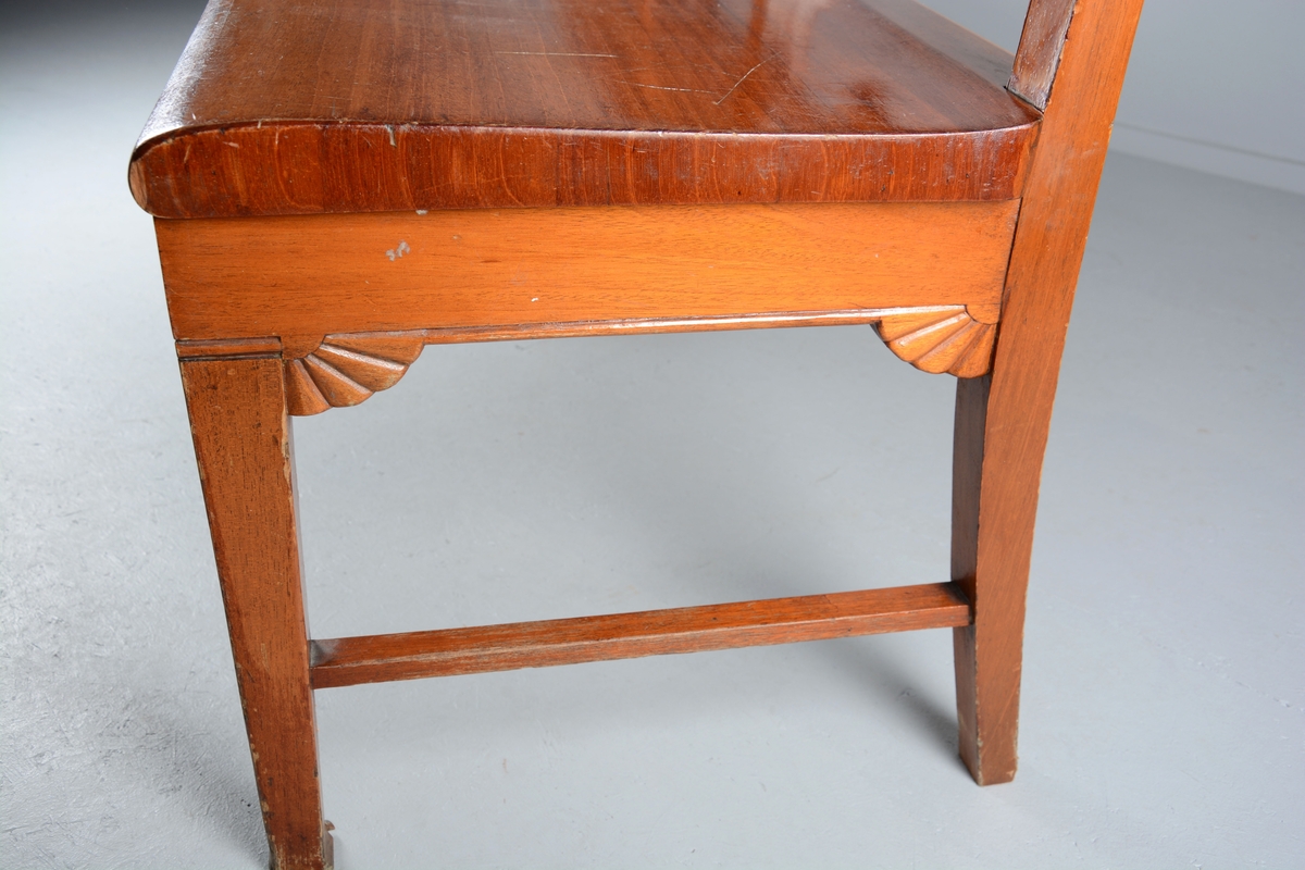 Sittebenk som har stått i Svaneapotekets kundeareal. Plass til 4 personer. Svakt buet ryggdel som danner ryggdel og ben på benken i samme stykke. Ryggen er delt i fire felt med lik dekor i hvert felt, 7 loddrette staver inni et rektangulært felt. En halvmåneformet blomst på midten av hvert felt på undersiden av det rektangulære feltet. Setet er laget i ett stykke tre og har en svak krumming oppover frem mot kanten på benken. Benken har tre sett med ben, to og to, på endene og på midten. Bena er parvis festet sammen med en sidesprosse. Rette framben. Under sargen på sidene og fremme er benken dekorert med samme type blomst som på ryggdelen, men her er de en halv halvmåne.