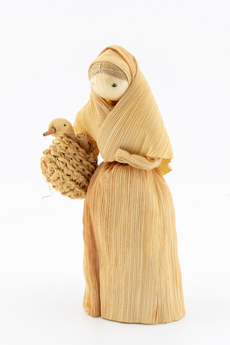 Pyntegjenstand i strå, forestiller kone i fotsid drakt med sjal over hodet, bærende på kurv med gås (?). Har tilhørt Sara Fabricius.