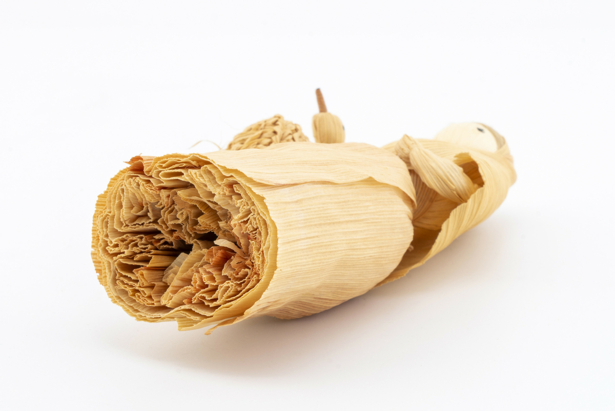 Pyntegjenstand i strå, forestiller kone i fotsid drakt med sjal over hodet, bærende på kurv med gås (?). Har tilhørt Sara Fabricius.