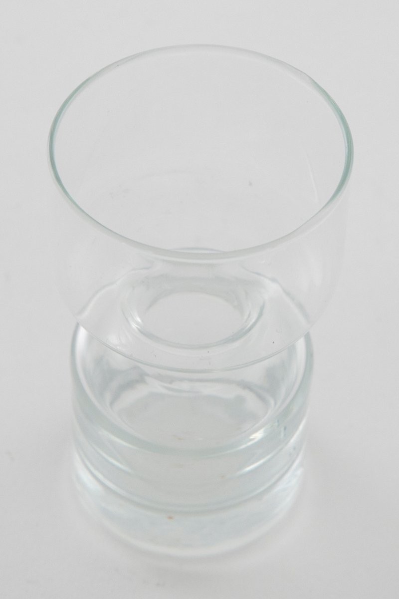 Lysestake i klart glass utformet som en sylinder som snevres inn på midten. Den øverste delen er litt videre enn den nederste.