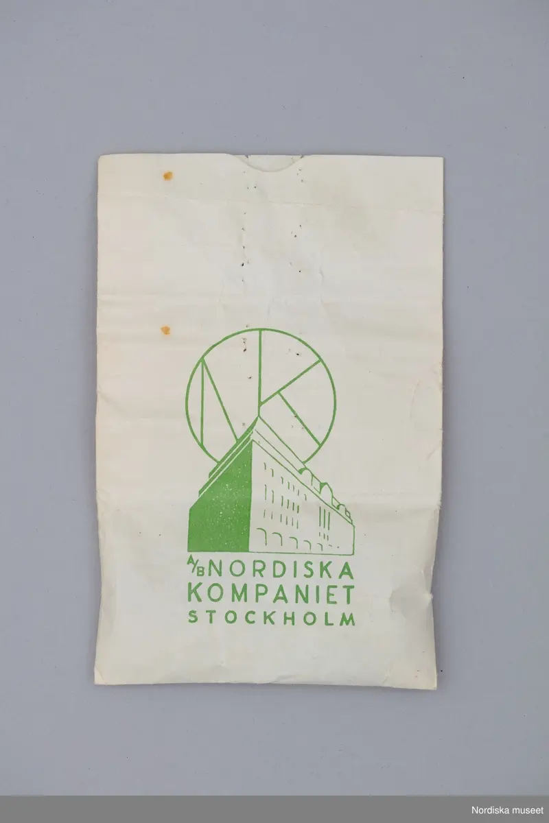 Arrangemang med rekvisitaföremål använda i samband med produktion av bok och utställning Nordens Paris. NK:s Franska damskrädderi 1902-1966. Här ses påse från NK, fingerborgar och glasstenar.