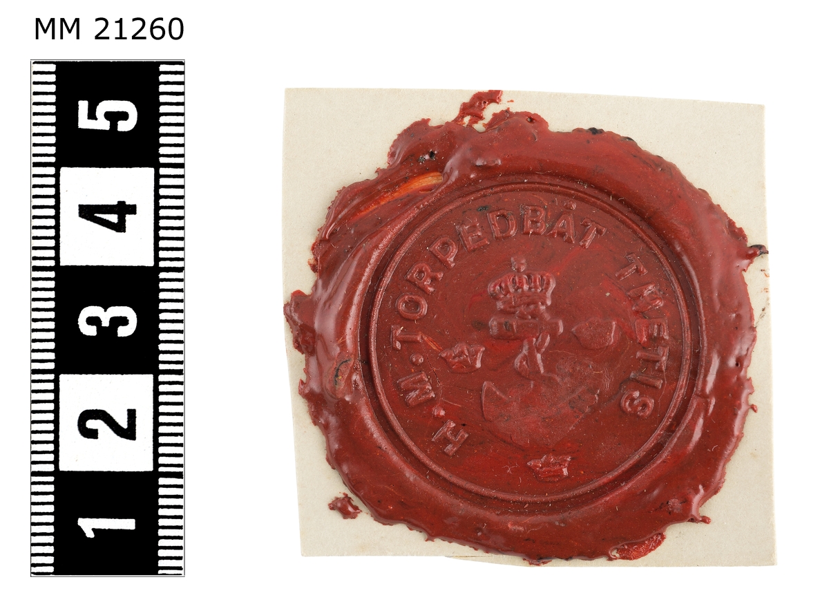 Sigillavtryck av rött lack på papper. I mitten krönt stockankare omgivet av tre kronor. Längs kanten text: "H.M. torpedbåt Thetis".