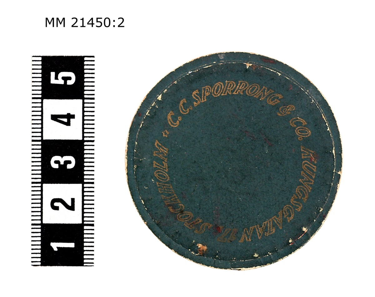 Ask av papper till minnespenning. Grönmålad utsida. På locket i guld "C.C. SPORRONG & CO KUNGSGATAN 17 STOCKHOLM".