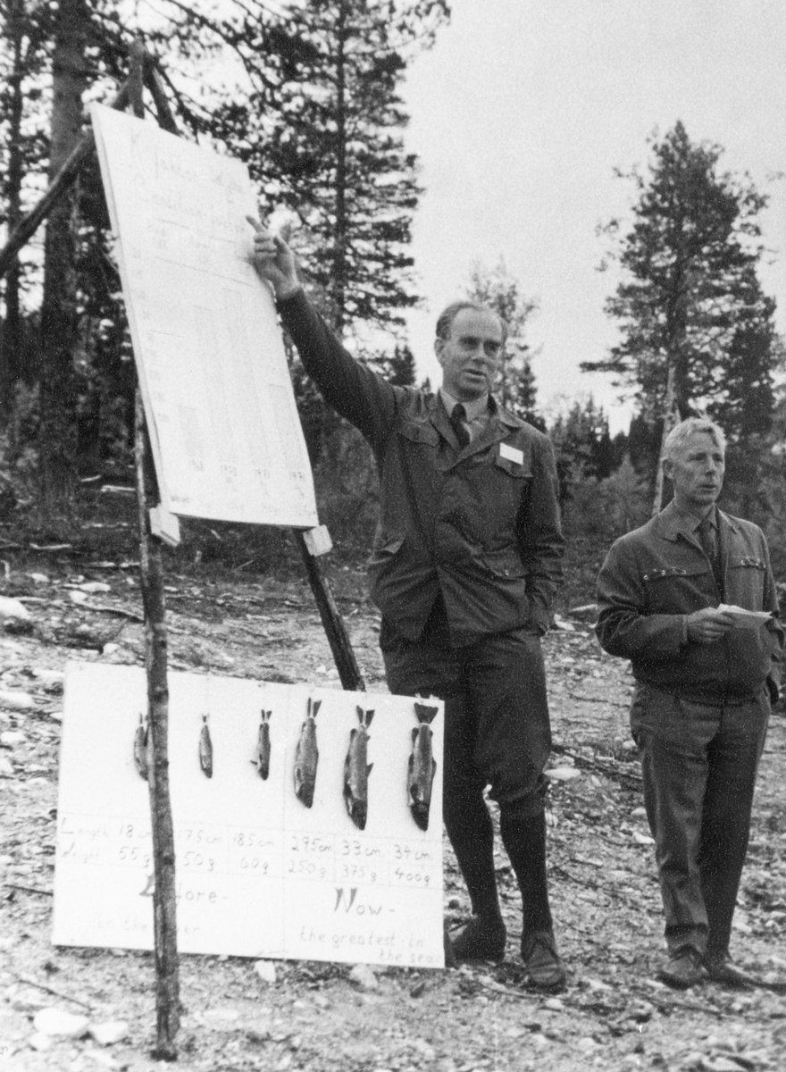 Skogdirektør Hans Kr. Seip orienterer om fisket ved Netfloen.  Orienteringa skjer på en åpen plass i skogen med furutrær i bakgrunnen.  Skogdirektøren har et stativ med tre bein, der han peker på et ark med søylediagrammer.  Underan står ei plansje med fisk av ulike størrelser. 