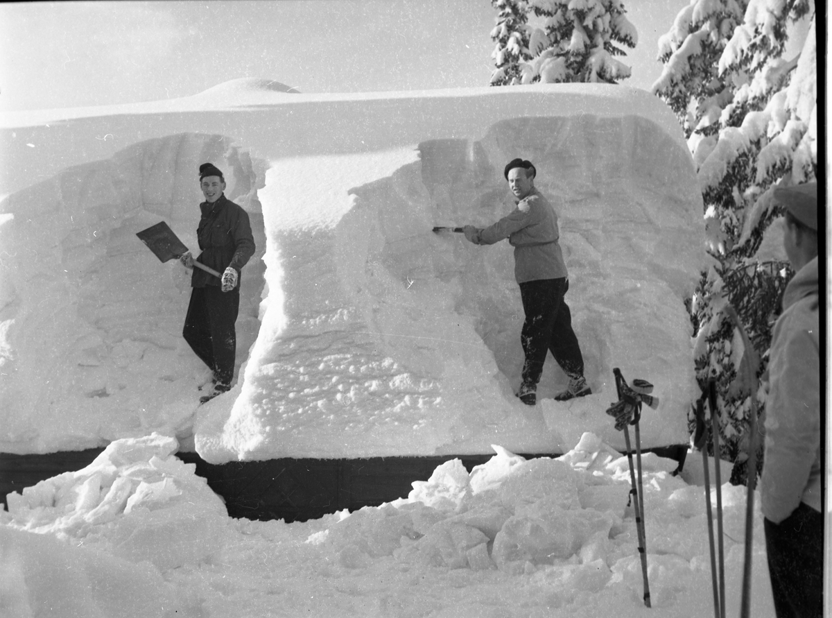 To menn jobber med å måke av et tak med mannshøy snø. En tredje står og ser på. 