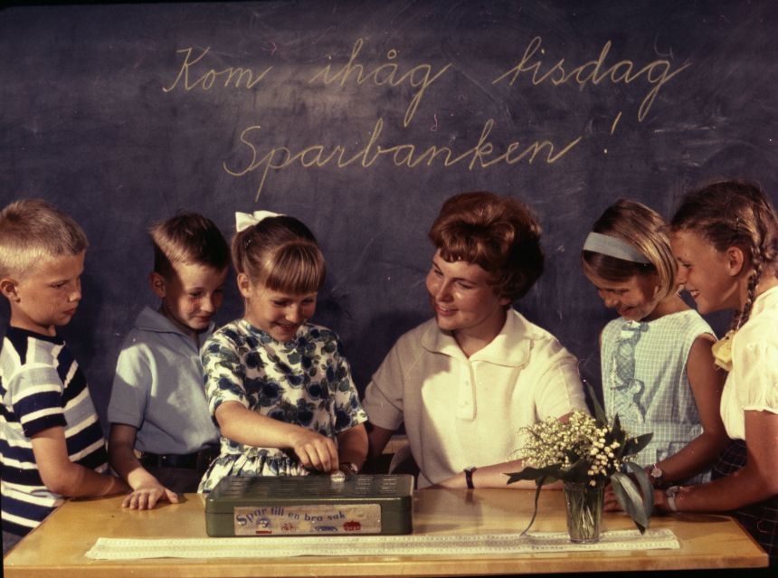 Skolsparsituation i klassrum. Barnen lägger sina pengar i
skolsparbössan under lärarinnans överinseende. På svarta tavlan står
uppmaningen. "Kom ihåg tisdag Sparbanken!"