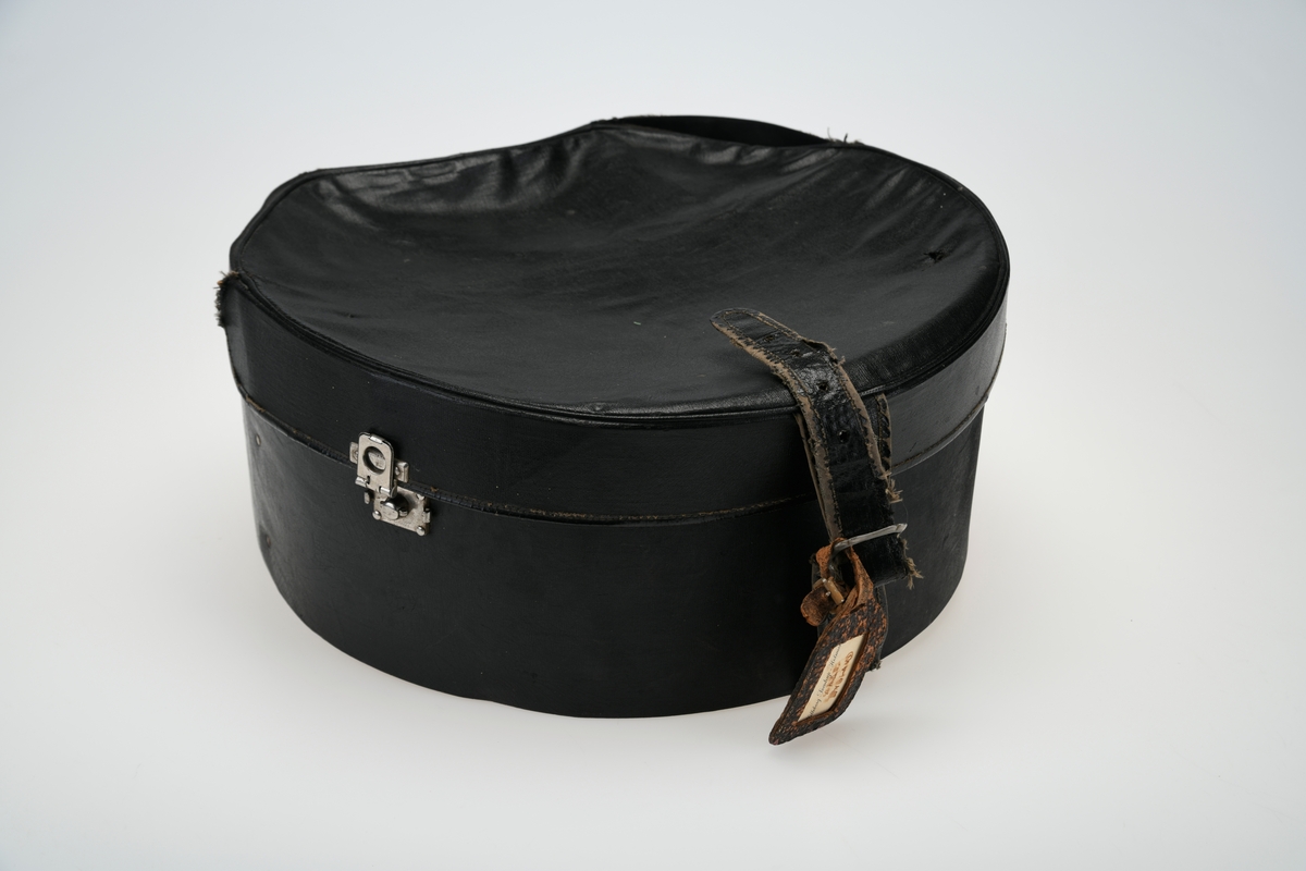 En hatteeske som i all hovedsak er rund men med avskåret bakende. Lokk og eske er laget av papp som er trukket med et plastbelagt lerretsstoff.
Kantene på lokk og eske er kantet med samme type tekstil/papir. I hver side av esken er det en lås av sølvfarget metall. De er naglet på. Låsemekanismen er rund og den ene delen er festet på esken mens den andre delen er festet på lokket. Den på lokket har fjærmekanisme og låses på den på esken. I front er det brede reimer med en spenne for å lukke/åpne. Spennen er avlang med avrundede hjørner og enkel dekor. Reimen som skal inni er bred innerst og smalner mot enden. Det er fem hull for stramming. På spennen er det festet en navnelapp av brunspraglet lær. På den står eiers/brukers navn og muligens detinasjon. Eksen er foret med svart linstoff. I hver side er det festet lange snorer av linstoff, trolig for å kunne "sikre" lasten i esken ved reise.