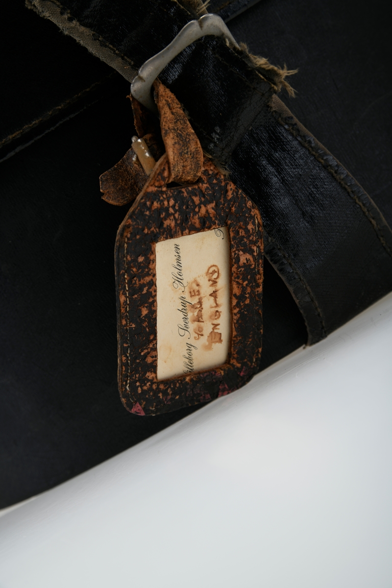 En hatteeske som i all hovedsak er rund men med avskåret bakende. Lokk og eske er laget av papp som er trukket med et plastbelagt lerretsstoff.
Kantene på lokk og eske er kantet med samme type tekstil/papir. I hver side av esken er det en lås av sølvfarget metall. De er naglet på. Låsemekanismen er rund og den ene delen er festet på esken mens den andre delen er festet på lokket. Den på lokket har fjærmekanisme og låses på den på esken. I front er det brede reimer med en spenne for å lukke/åpne. Spennen er avlang med avrundede hjørner og enkel dekor. Reimen som skal inni er bred innerst og smalner mot enden. Det er fem hull for stramming. På spennen er det festet en navnelapp av brunspraglet lær. På den står eiers/brukers navn og muligens detinasjon. Eksen er foret med svart linstoff. I hver side er det festet lange snorer av linstoff, trolig for å kunne "sikre" lasten i esken ved reise.