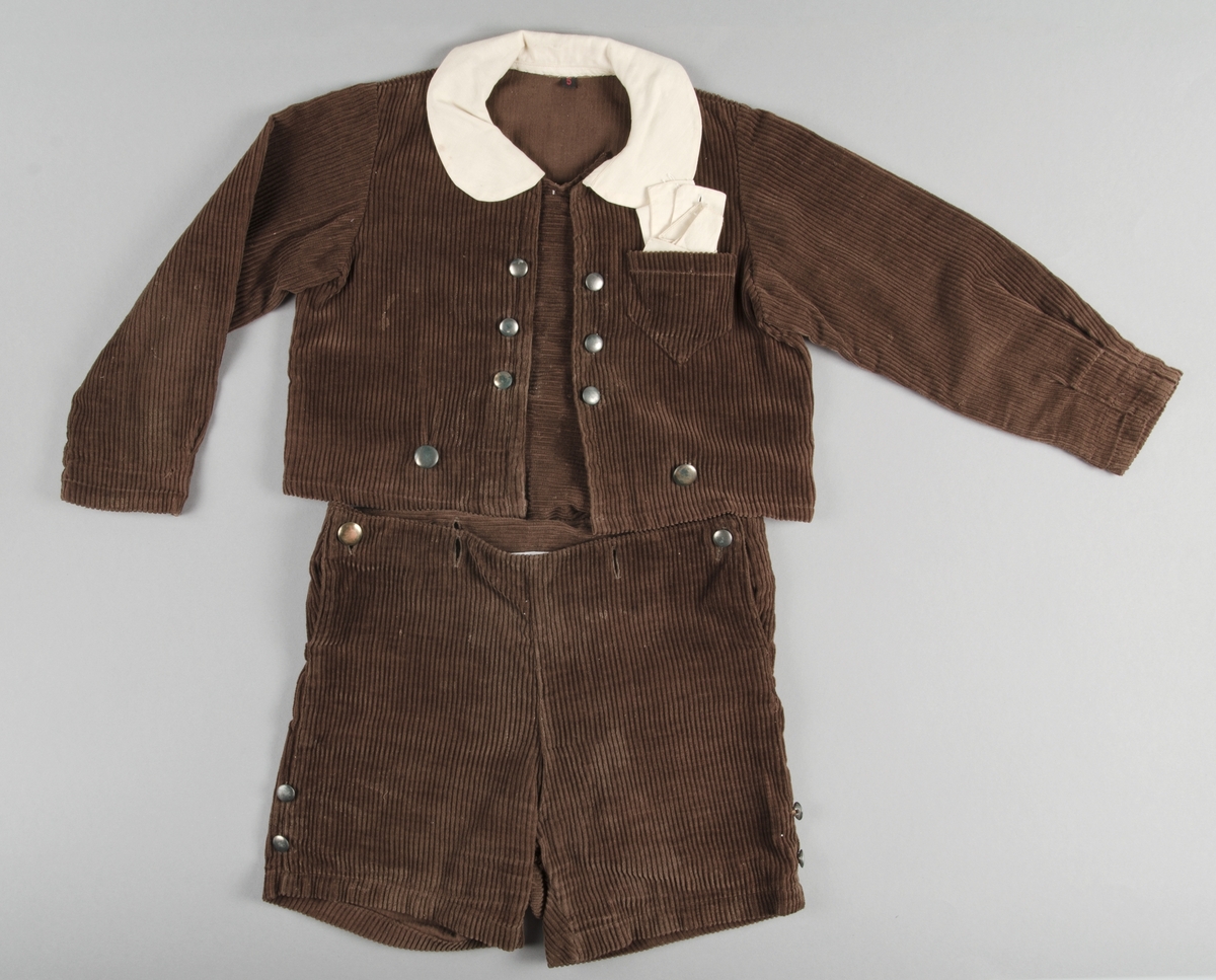 Dräkt för pojke ca 3-5 år. 1920-30-tal. Jacka och kortbyxor i brun manschester. Jackan har lång ärm, knäppes fram med sex blanka metallknappar. I nederkanten av jackan finns ytterligare knappar att fästa byxorna vid. Dräktjackan har löstagbara manchetter, krage och näsduk av gulnat bomulltyg. Bröstficka för näsduk på vänster sida. 
Kortbyxorna knäppes i sidan med runda metallknappar. Knapphål i byxlinningen för för att fästa jackan.

