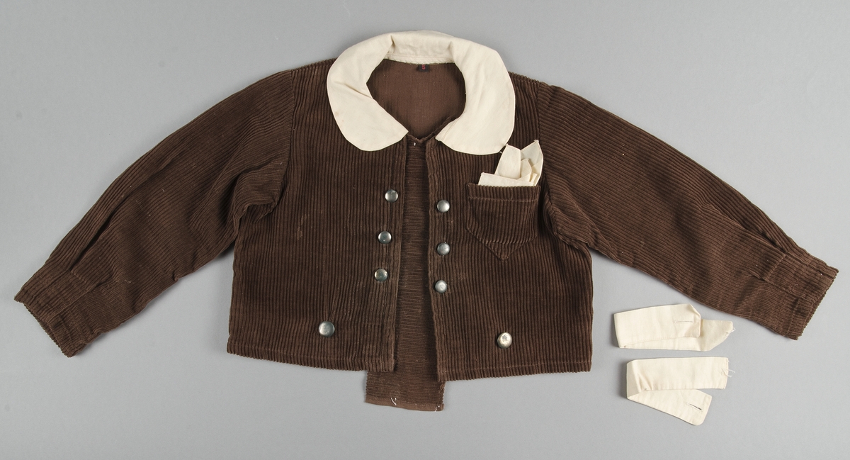 Dräkt för pojke ca 3-5 år. 1920-30-tal. Jacka och kortbyxor i brun manschester. Jackan har lång ärm, knäppes fram med sex blanka metallknappar. I nederkanten av jackan finns ytterligare knappar att fästa byxorna vid. Dräktjackan har löstagbara manchetter, krage och näsduk av gulnat bomulltyg. Bröstficka för näsduk på vänster sida. 
Kortbyxorna knäppes i sidan med runda metallknappar. Knapphål i byxlinningen för för att fästa jackan.

