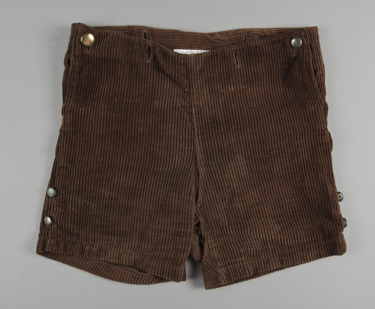 Dräkt för pojke ca 3-5 år. 1920-30-tal. Jacka och kortbyxor i brun manschester. Jackan har lång ärm, knäppes fram med sex blanka metallknappar. I nederkanten av jackan finns ytterligare knappar att fästa byxorna vid. Dräktjackan har löstagbara manchetter, krage och näsduk av gulnat bomulltyg. Bröstficka för näsduk på vänster sida. 
Kortbyxorna knäppes i sidan med runda metallknappar. Knapphål i byxlinningen för för att fästa jackan.

