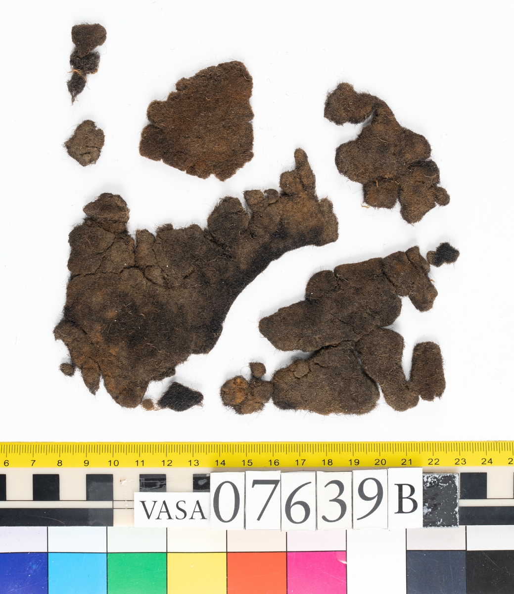 Textilfragment. 53 fragment fördelade på fyndnummer 07639a-e.
Fnr 07639a består av 17 fragment av ull vävda i tuskaft.
Fnr 07639b består av 8 fragment av hattfilt.
Fnr 07639c består av 17 fragment av ull vävda i kypert.
Fnr 07639d består av 1 fragment av ull vävt i kypert.
Fnr 07639e har 10 fragment av ull vävda i tuskaft varav ett fragment har en stadkant.

Textildokumentation 2017:
07639b
9 fragment i hårt filtad filt. Filten är tjock och kompakt, typ 1:1 har även en klippt eller skuren del. Sidan ett (synlig på bilden 1) är mycket mörkare i ytan, nästan svart, medan sida två går mer åt brunt. Båda sidorna har synliga märken efter avtryck, På sida två syns dessa bäst. En del hår i filten är finare än andra och dessa blänker mer. Möjligen är materialet bäver.
07639d:
Ett tygfragment i kompakt, tät och grov kypert, typ 3:1. Fragmentet har kraftigt upprepade kanter runtom. Tjocka fransar har bildats på den längsta sidan, dessa består av trådsystem 1. Trådsystem 1 är mycket hårt Z-tvinnat, nästan så att det lägger sig i öglor. Tyget är glansigt och på sida ett ser det något ripsartat ut, då trådsystem 1 nästan inte syns, utan bara randningen.  Sida två av tyget har nötningar och glesare kypertränder.