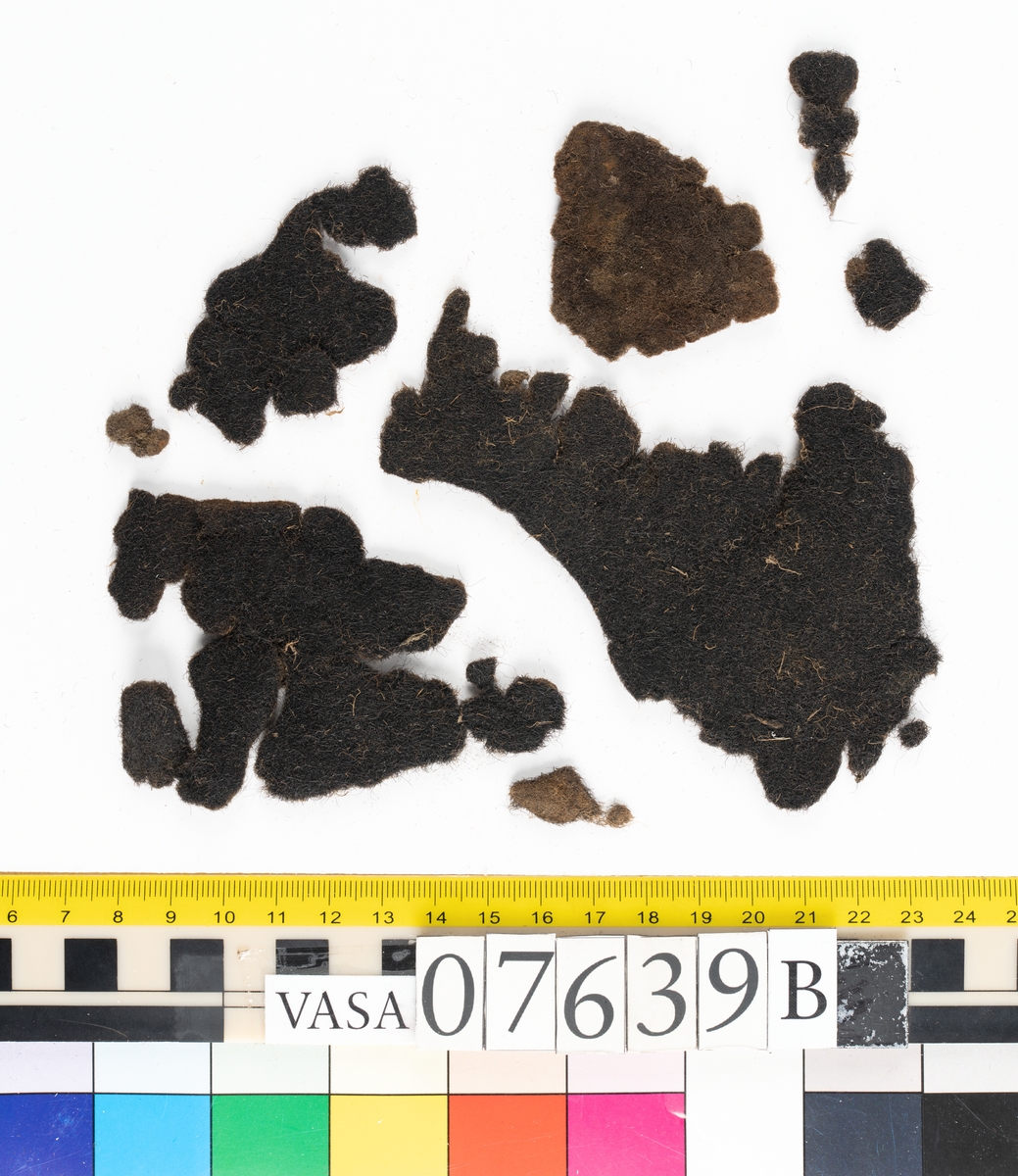 Textilfragment. 53 fragment fördelade på fyndnummer 07639a-e.
Fnr 07639a består av 17 fragment av ull vävda i tuskaft.
Fnr 07639b består av 8 fragment av hattfilt.
Fnr 07639c består av 17 fragment av ull vävda i kypert.
Fnr 07639d består av 1 fragment av ull vävt i kypert.
Fnr 07639e har 10 fragment av ull vävda i tuskaft varav ett fragment har en stadkant.

Textildokumentation 2017:
07639b
9 fragment i hårt filtad filt. Filten är tjock och kompakt, typ 1:1 har även en klippt eller skuren del. Sidan ett (synlig på bilden 1) är mycket mörkare i ytan, nästan svart, medan sida två går mer åt brunt. Båda sidorna har synliga märken efter avtryck, På sida två syns dessa bäst. En del hår i filten är finare än andra och dessa blänker mer. Möjligen är materialet bäver.
07639d:
Ett tygfragment i kompakt, tät och grov kypert, typ 3:1. Fragmentet har kraftigt upprepade kanter runtom. Tjocka fransar har bildats på den längsta sidan, dessa består av trådsystem 1. Trådsystem 1 är mycket hårt Z-tvinnat, nästan så att det lägger sig i öglor. Tyget är glansigt och på sida ett ser det något ripsartat ut, då trådsystem 1 nästan inte syns, utan bara randningen.  Sida två av tyget har nötningar och glesare kypertränder.