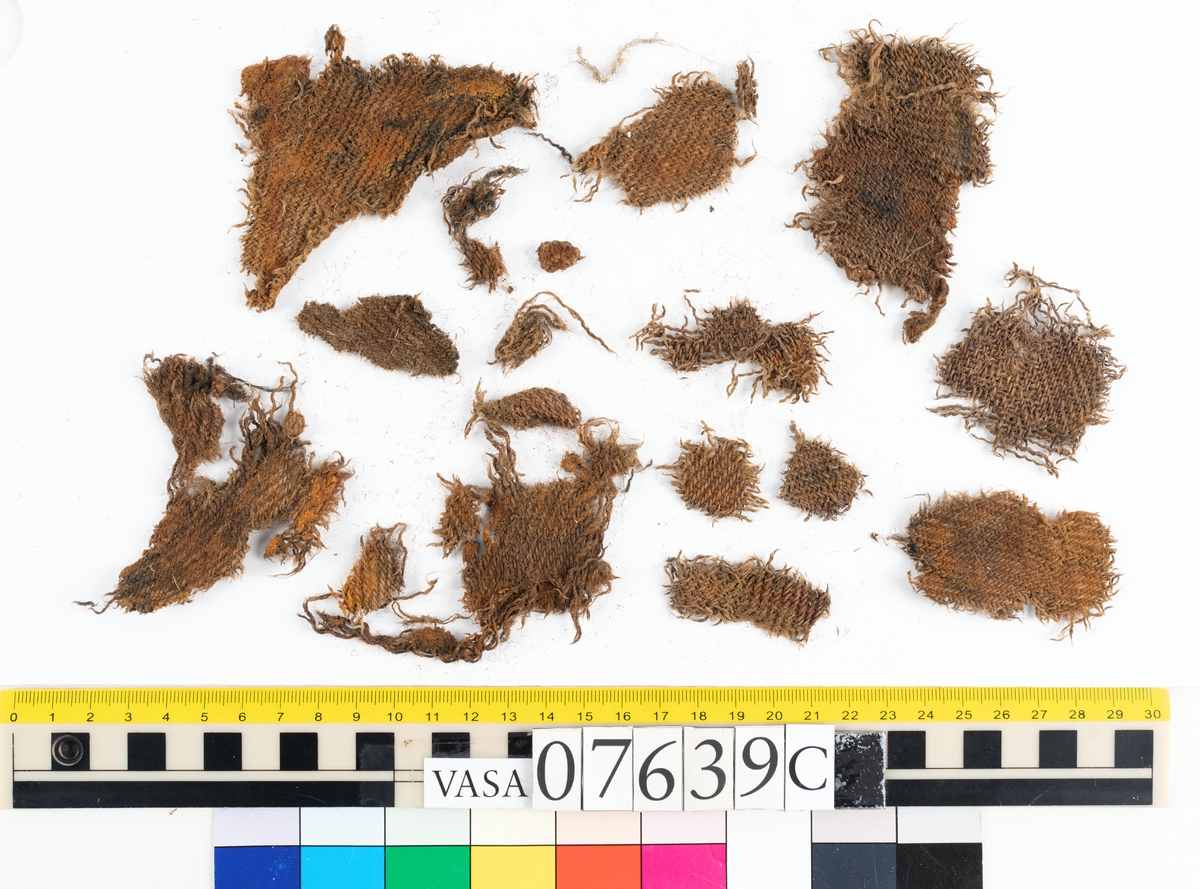 Textilfragment. 53 fragment fördelade på fyndnummer 07639a-e.
Fnr 07639a består av 17 fragment av ull vävda i tuskaft.
Fnr 07639b består av 8 fragment av hattfilt.
Fnr 07639c består av 17 fragment av ull vävda i kypert.
Fnr 07639d består av 1 fragment av ull vävt i kypert.
Fnr 07639e har 10 fragment av ull vävda i tuskaft varav ett fragment har en stadkant.

Textildokumentation 2017:
07639b
9 fragment i hårt filtad filt. Filten är tjock och kompakt, typ 1:1 har även en klippt eller skuren del. Sidan ett (synlig på bilden 1) är mycket mörkare i ytan, nästan svart, medan sida två går mer åt brunt. Båda sidorna har synliga märken efter avtryck, På sida två syns dessa bäst. En del hår i filten är finare än andra och dessa blänker mer. Möjligen är materialet bäver.
07639d:
Ett tygfragment i kompakt, tät och grov kypert, typ 3:1. Fragmentet har kraftigt upprepade kanter runtom. Tjocka fransar har bildats på den längsta sidan, dessa består av trådsystem 1. Trådsystem 1 är mycket hårt Z-tvinnat, nästan så att det lägger sig i öglor. Tyget är glansigt och på sida ett ser det något ripsartat ut, då trådsystem 1 nästan inte syns, utan bara randningen.  Sida två av tyget har nötningar och glesare kypertränder.