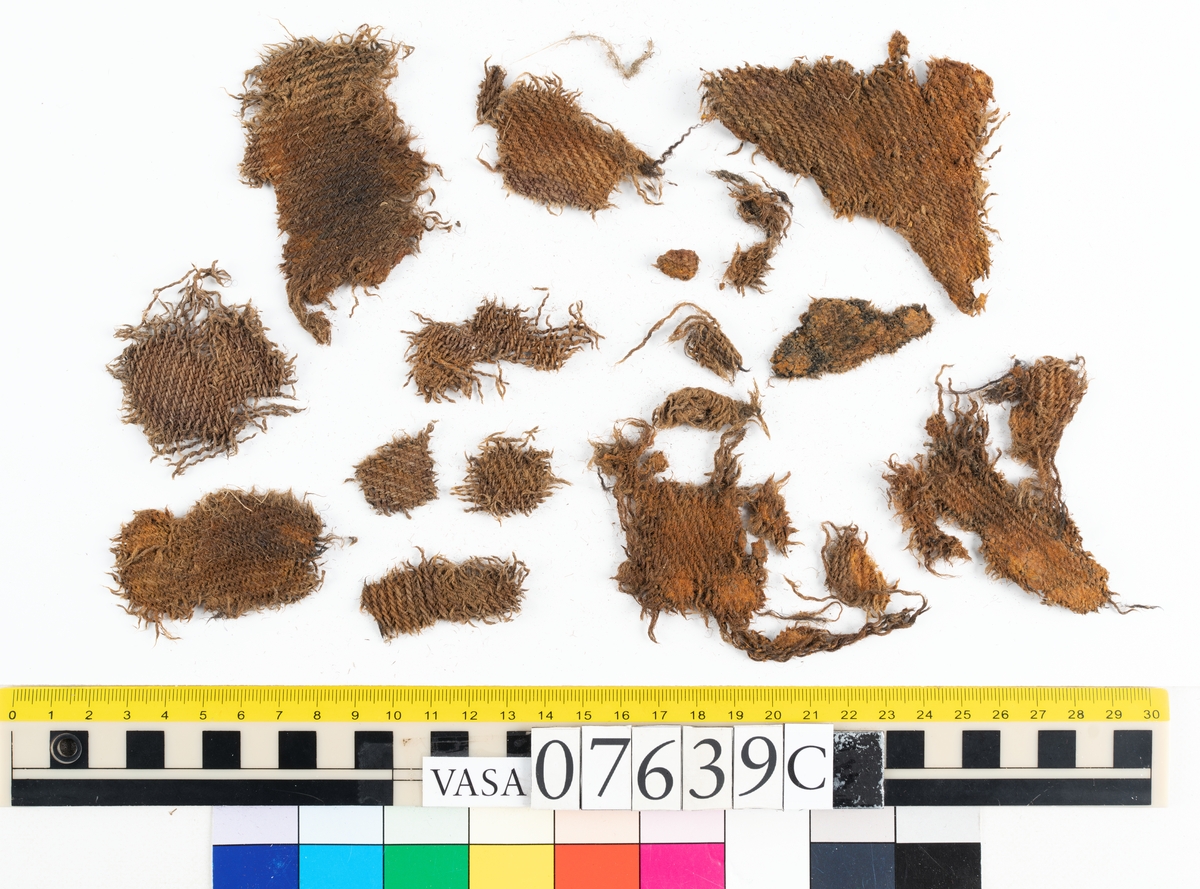 Textilfragment. 53 fragment fördelade på fyndnummer 07639a-e.
Fnr 07639a består av 17 fragment av ull vävda i tuskaft.
Fnr 07639b består av 8 fragment av hattfilt.
Fnr 07639c består av 17 fragment av ull vävda i kypert.
Fnr 07639d består av 1 fragment av ull vävt i kypert.
Fnr 07639e har 10 fragment av ull vävda i tuskaft varav ett fragment har en stadkant.

Textildokumentation 2017:
07639b
9 fragment i hårt filtad filt. Filten är tjock och kompakt, typ 1:1 har även en klippt eller skuren del. Sidan ett (synlig på bilden 1) är mycket mörkare i ytan, nästan svart, medan sida två går mer åt brunt. Båda sidorna har synliga märken efter avtryck, På sida två syns dessa bäst. En del hår i filten är finare än andra och dessa blänker mer. Möjligen är materialet bäver.
07639d:
Ett tygfragment i kompakt, tät och grov kypert, typ 3:1. Fragmentet har kraftigt upprepade kanter runtom. Tjocka fransar har bildats på den längsta sidan, dessa består av trådsystem 1. Trådsystem 1 är mycket hårt Z-tvinnat, nästan så att det lägger sig i öglor. Tyget är glansigt och på sida ett ser det något ripsartat ut, då trådsystem 1 nästan inte syns, utan bara randningen.  Sida två av tyget har nötningar och glesare kypertränder.