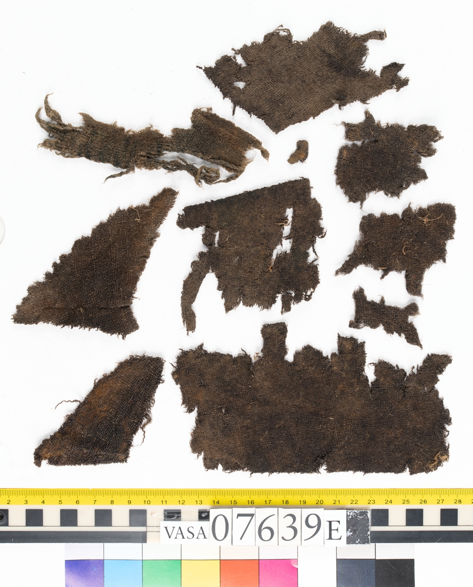 Textilfragment. 53 fragment fördelade på fyndnummer 07639a-e.
Fnr 07639a består av 17 fragment av ull vävda i tuskaft.
Fnr 07639b består av 8 fragment av hattfilt.
Fnr 07639c består av 17 fragment av ull vävda i kypert.
Fnr 07639d består av 1 fragment av ull vävt i kypert.
Fnr 07639e har 10 fragment av ull vävda i tuskaft varav ett fragment har en stadkant.

Textildokumentation 2017:
07639b
9 fragment i hårt filtad filt. Filten är tjock och kompakt, typ 1:1 har även en klippt eller skuren del. Sidan ett (synlig på bilden 1) är mycket mörkare i ytan, nästan svart, medan sida två går mer åt brunt. Båda sidorna har synliga märken efter avtryck, På sida två syns dessa bäst. En del hår i filten är finare än andra och dessa blänker mer. Möjligen är materialet bäver.
07639d:
Ett tygfragment i kompakt, tät och grov kypert, typ 3:1. Fragmentet har kraftigt upprepade kanter runtom. Tjocka fransar har bildats på den längsta sidan, dessa består av trådsystem 1. Trådsystem 1 är mycket hårt Z-tvinnat, nästan så att det lägger sig i öglor. Tyget är glansigt och på sida ett ser det något ripsartat ut, då trådsystem 1 nästan inte syns, utan bara randningen.  Sida två av tyget har nötningar och glesare kypertränder.