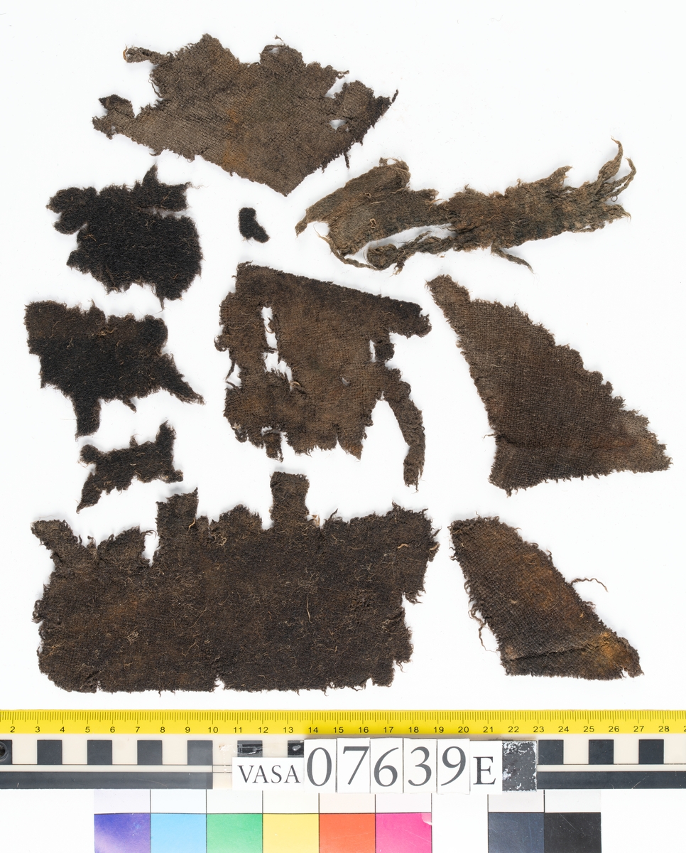 Textilfragment. 53 fragment fördelade på fyndnummer 07639a-e.
Fnr 07639a består av 17 fragment av ull vävda i tuskaft.
Fnr 07639b består av 8 fragment av hattfilt.
Fnr 07639c består av 17 fragment av ull vävda i kypert.
Fnr 07639d består av 1 fragment av ull vävt i kypert.
Fnr 07639e har 10 fragment av ull vävda i tuskaft varav ett fragment har en stadkant.

Textildokumentation 2017:
07639b
9 fragment i hårt filtad filt. Filten är tjock och kompakt, typ 1:1 har även en klippt eller skuren del. Sidan ett (synlig på bilden 1) är mycket mörkare i ytan, nästan svart, medan sida två går mer åt brunt. Båda sidorna har synliga märken efter avtryck, På sida två syns dessa bäst. En del hår i filten är finare än andra och dessa blänker mer. Möjligen är materialet bäver.
07639d:
Ett tygfragment i kompakt, tät och grov kypert, typ 3:1. Fragmentet har kraftigt upprepade kanter runtom. Tjocka fransar har bildats på den längsta sidan, dessa består av trådsystem 1. Trådsystem 1 är mycket hårt Z-tvinnat, nästan så att det lägger sig i öglor. Tyget är glansigt och på sida ett ser det något ripsartat ut, då trådsystem 1 nästan inte syns, utan bara randningen.  Sida två av tyget har nötningar och glesare kypertränder.