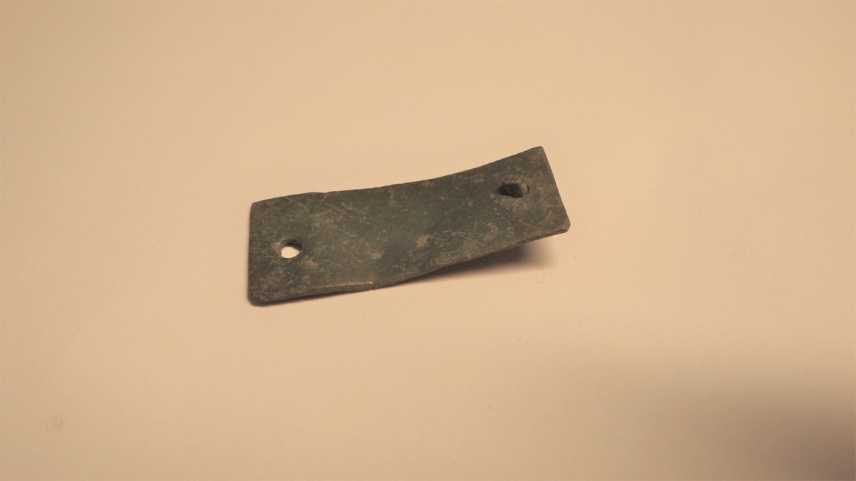 Rektangulær metallplate med to naglehull, et i hver ende. I det ene hullet sitter en nagle.