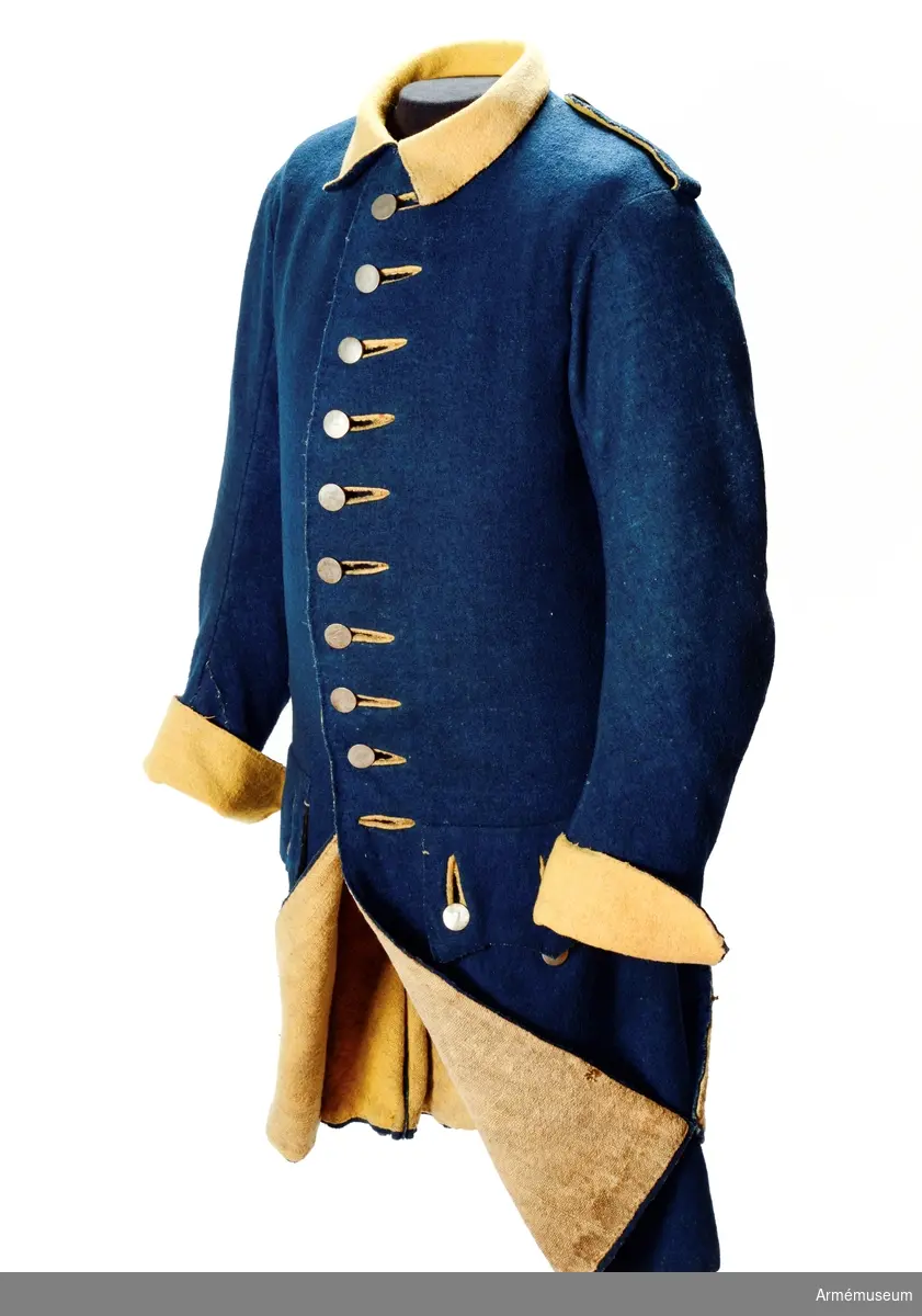 Rock av blått kläde med gul krage och ärmuppslag, fodrad med gul boj i livet samt linne i ärmarna. Besatt med tennknappar. Märke efter lacksigill.