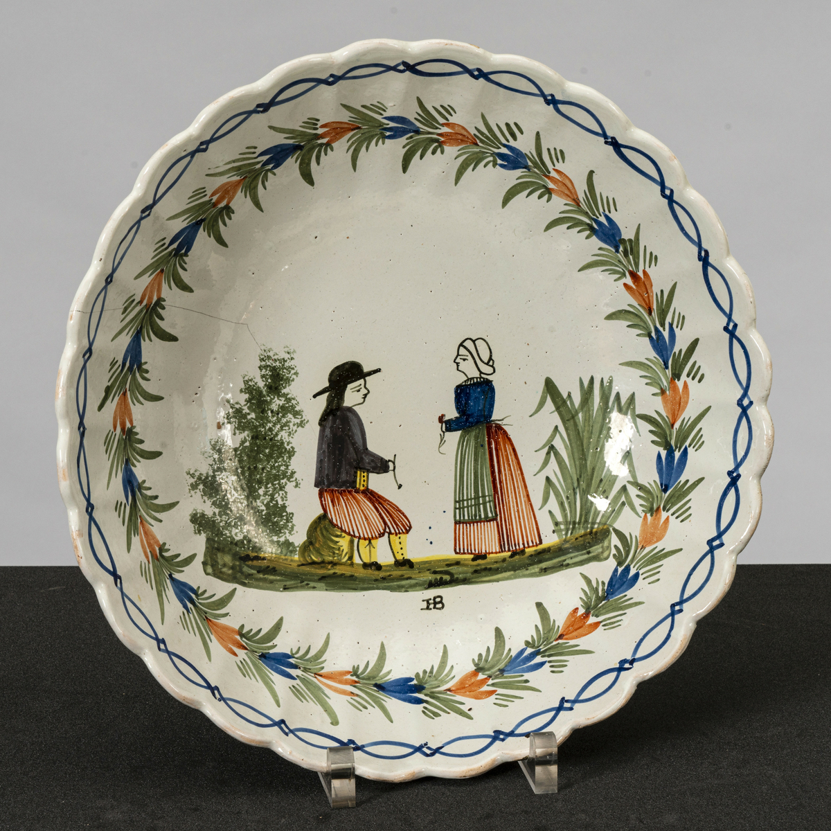 Rundt, dypt, stort serveringsfat med hull til oppheng på basen. Bølget kant og riller i kanten mot toppen. Påmalt bord mot kanten, blad-/ blomsterranke under og motiv av kvinne og mann i tradisjonelle drakter (stakk, forkle, nikkers, vest, hatter) vendt mot hverandre midt på bunnen. Mannen sitter på en stein, kvinnen står, begge holder en blomst. Har tilhørt Sara Fabricius.