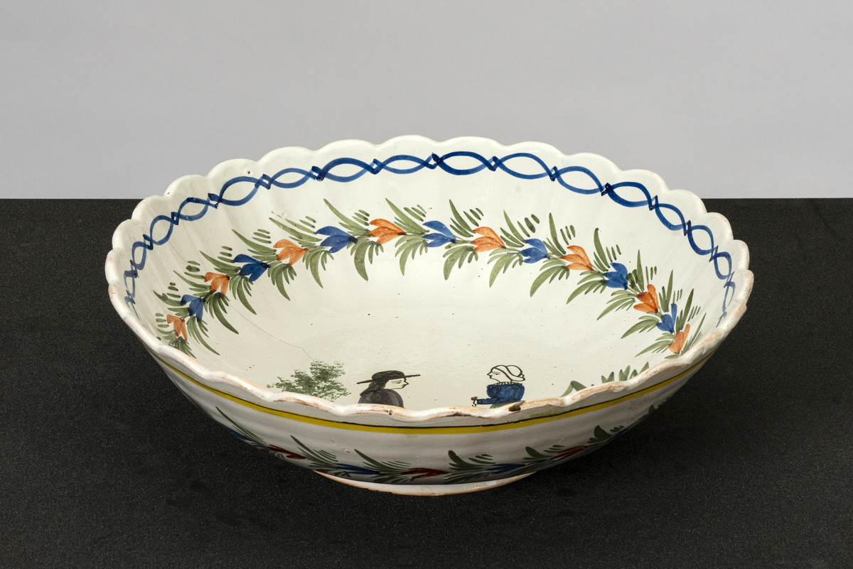 Rundt, dypt, stort serveringsfat med hull til oppheng på basen. Bølget kant og riller i kanten mot toppen. Påmalt bord mot kanten, blad-/ blomsterranke under og motiv av kvinne og mann i tradisjonelle drakter (stakk, forkle, nikkers, vest, hatter) vendt mot hverandre midt på bunnen. Mannen sitter på en stein, kvinnen står, begge holder en blomst. Har tilhørt Sara Fabricius.