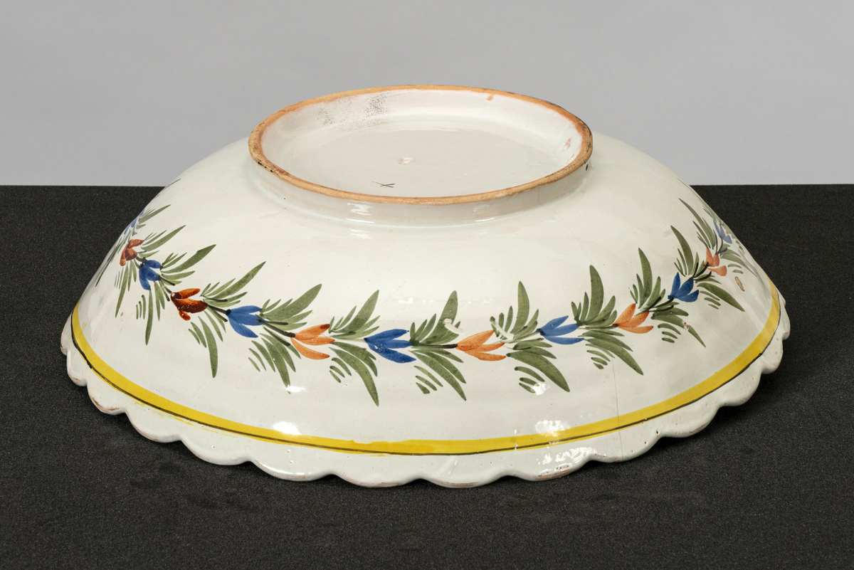 Rundt, dypt, stort serveringsfat med hull til oppheng på basen. Bølget kant og riller i kanten mot toppen. Påmalt bord mot kanten, blad-/ blomsterranke under og motiv av kvinne og mann i tradisjonelle drakter (stakk, forkle, nikkers, vest, hatter) vendt mot hverandre midt på bunnen. Mannen sitter på en stein, kvinnen står, begge holder en blomst. Har tilhørt Sara Fabricius.