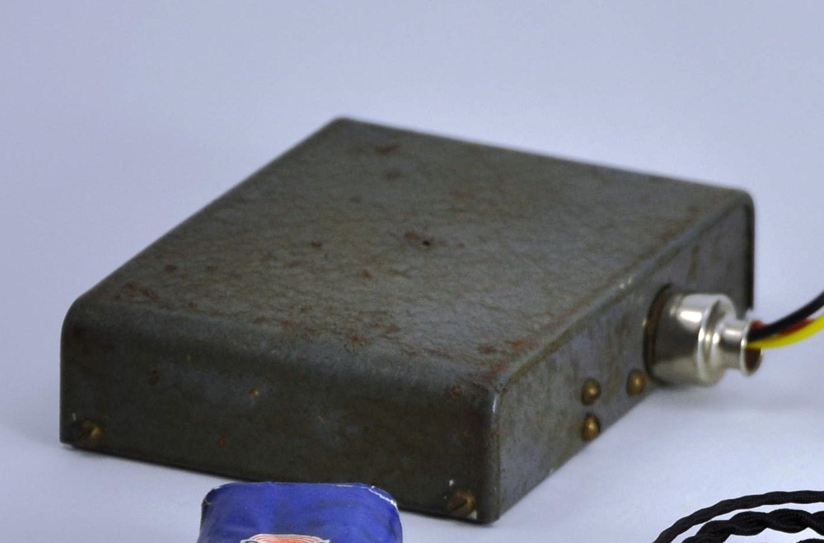 Batteriboks i samme farge, materiale og fasong som radioen, men mindre. På den ene langsiden er uttaket til batteripluggen.