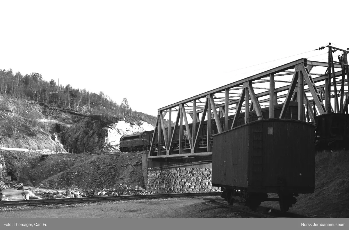 En av Sulitjelmabanens flisvogner litra G nr. 104 eller 103 på Finneid stasjon. I bakgrunnen diesellokomotiv Di 3 med godstog på Finneidstrømmen bru