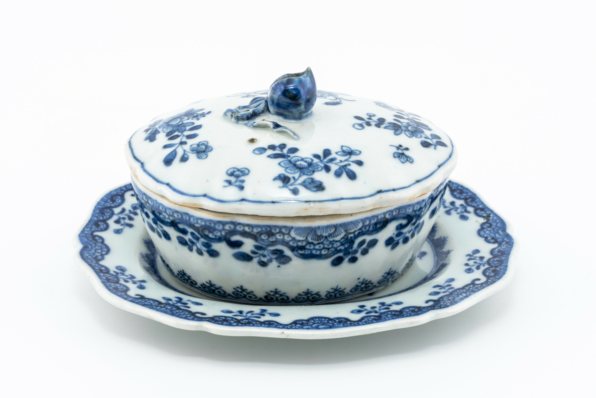 Oval keramikkskål med lokk, på fat. Hvit med påmalt blå blomsterdekor. Grep i påmontert porselen i form av en blomsterknopp. Inneholder en liten terning i bein. Har tilhørt Sara Fabricius.