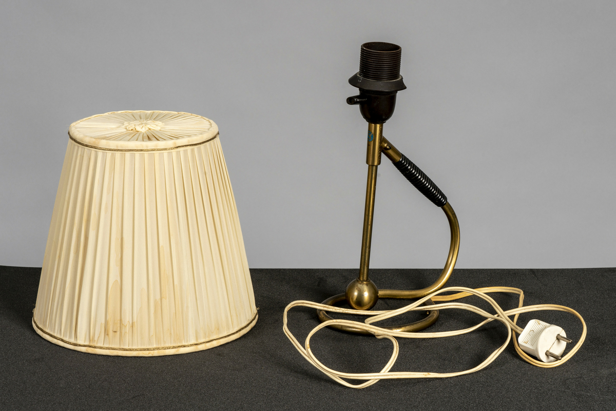 Bordlampe i messing med håndtak i plast. Tilhørende plissert kremfarget tekstilskjerm med 6 spiler, dekorbånd i gullfarge og tekstilblomst på toppen. Av/på-vrider på pæresokkel. Har tilhørt Sara Fabricius.