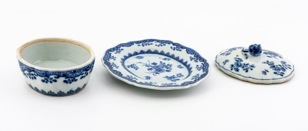 Oval keramikkskål med lokk, på fat. Hvit med påmalt blå blomsterdekor. Grep i påmontert porselen i form av en blomsterknopp. Inneholder en liten terning i bein. Har tilhørt Sara Fabricius.