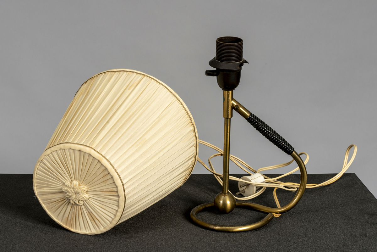 Bordlampe i messing med håndtak i plast. Tilhørende plissert kremfarget tekstilskjerm med 6 spiler, dekorbånd i gullfarge og tekstilblomst på toppen. Av/på-vrider på pæresokkel. Har tilhørt Sara Fabricius.