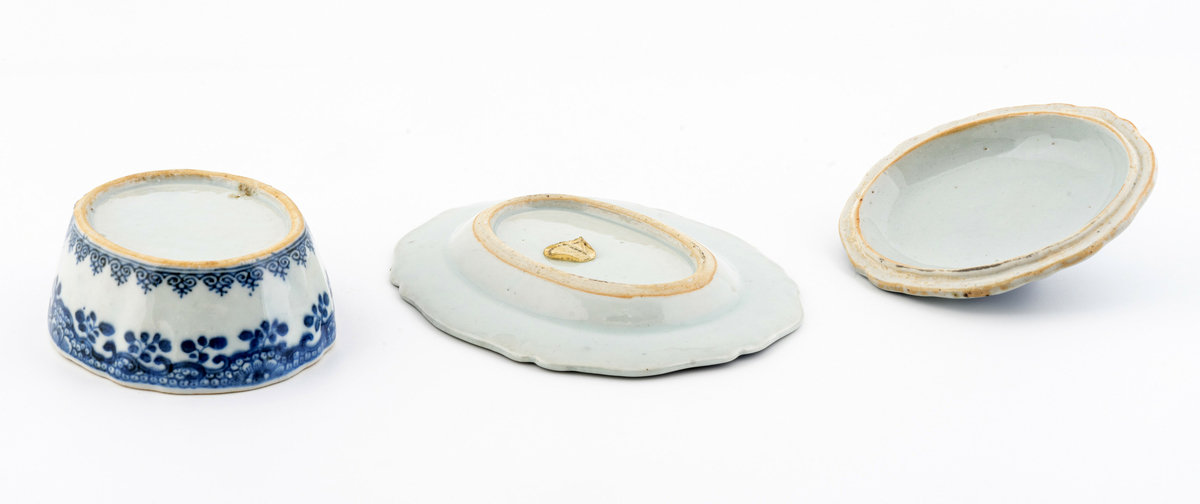 Oval keramikkskål med lokk, på fat. Hvit med påmalt blå blomsterdekor. Grep i påmontert porselen i form av en blomsterknopp. Inneholder en liten terning i bein. Har tilhørt Sara Fabricius.