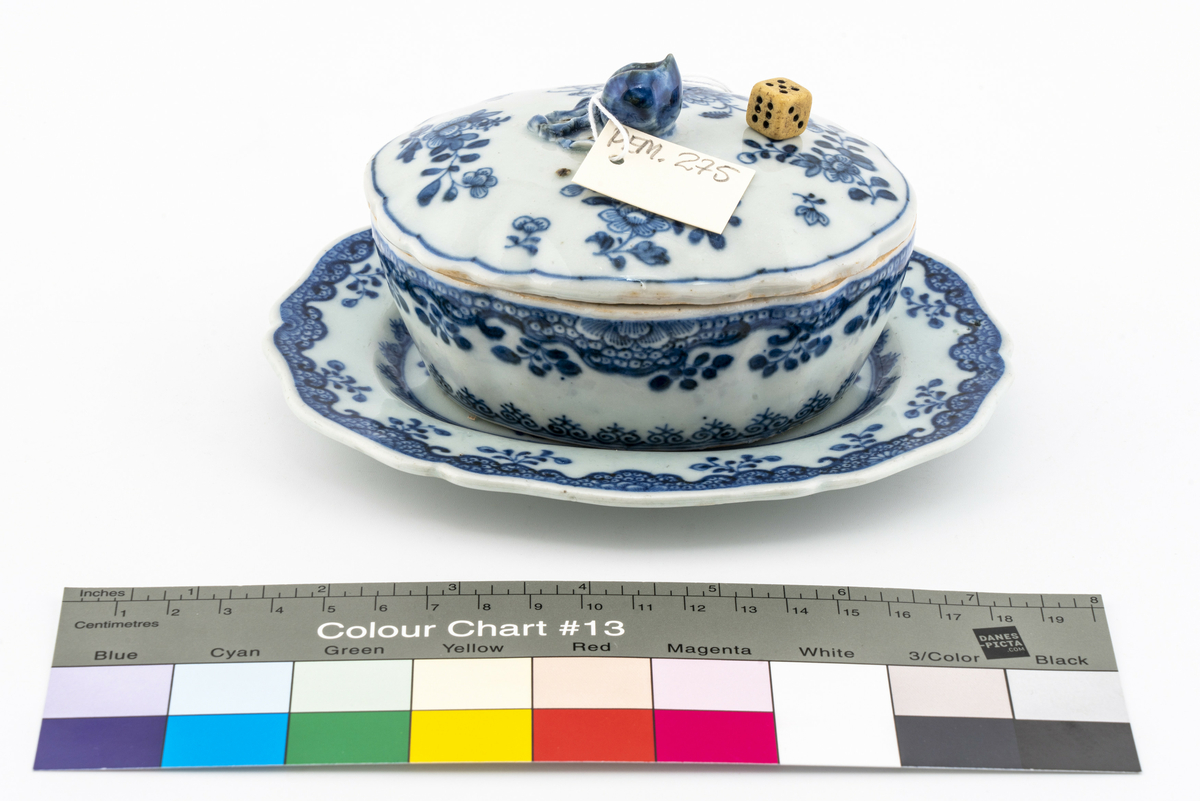 Oval keramikkskål med lokk, på fat. Hvit med påmalt blå blomsterdekor. Grep i påmontert porselen i form av en blomsterknopp. Inneholder en liten terning i bein. Har tilhørt Sara Fabricius.