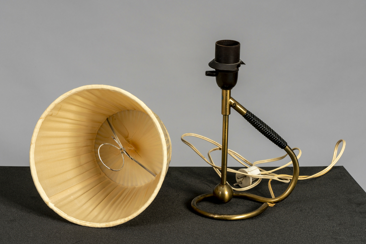 Bordlampe i messing med håndtak i plast. Tilhørende plissert kremfarget tekstilskjerm med 6 spiler, dekorbånd i gullfarge og tekstilblomst på toppen. Av/på-vrider på pæresokkel. Har tilhørt Sara Fabricius.