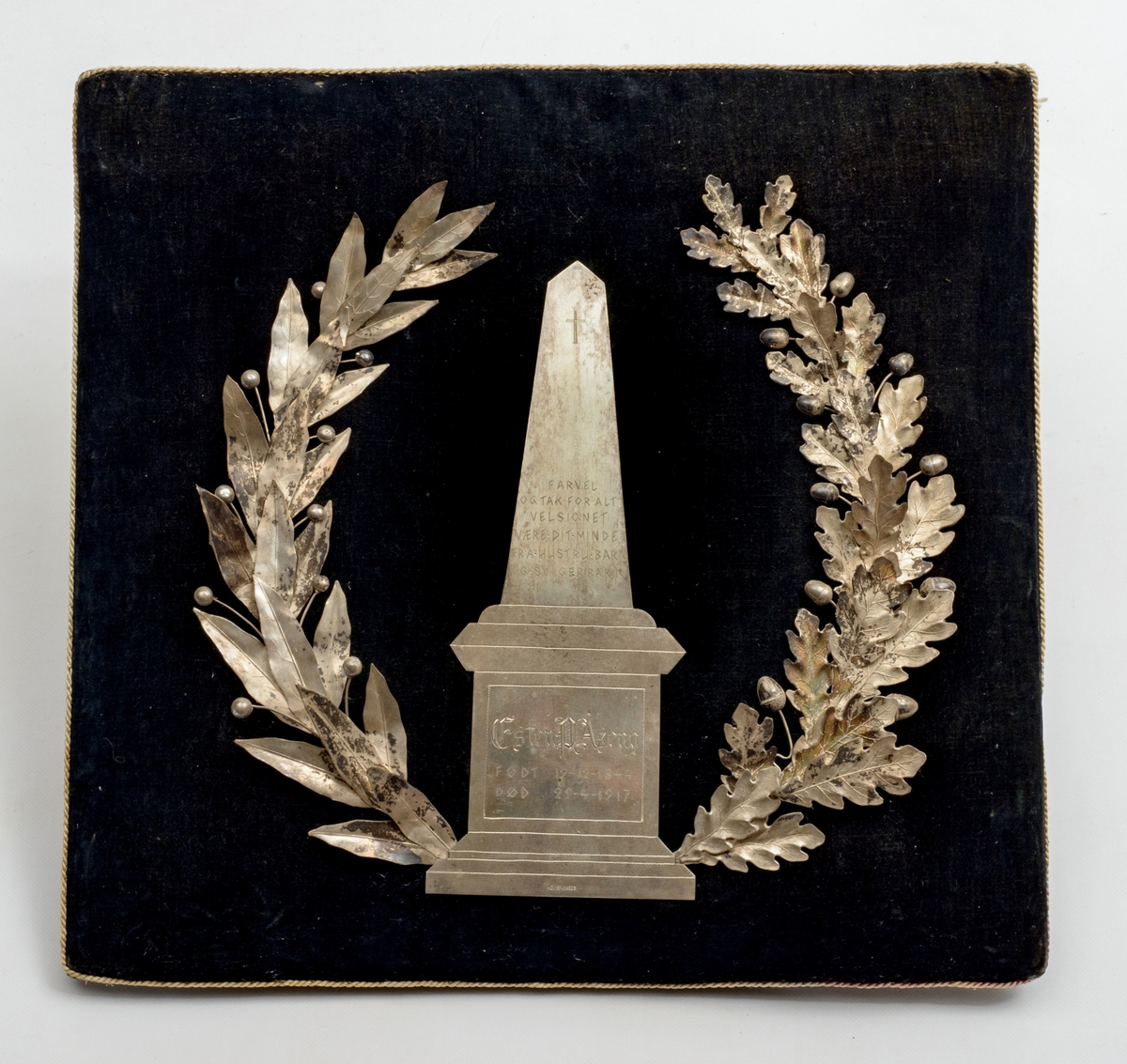 Krans på pute. Den ene delen av kransen har eikeblad og eikenøtter som motiv, og den andre delen har en flatere type blad med knopper. I midten er det en obelisk på en plattform.
