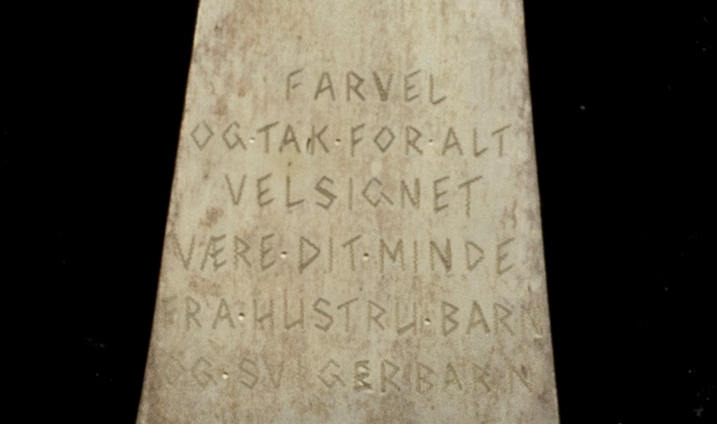Krans på pute. Den ene delen av kransen har eikeblad og eikenøtter som motiv, og den andre delen har en flatere type blad med knopper. I midten er det en obelisk på en plattform.